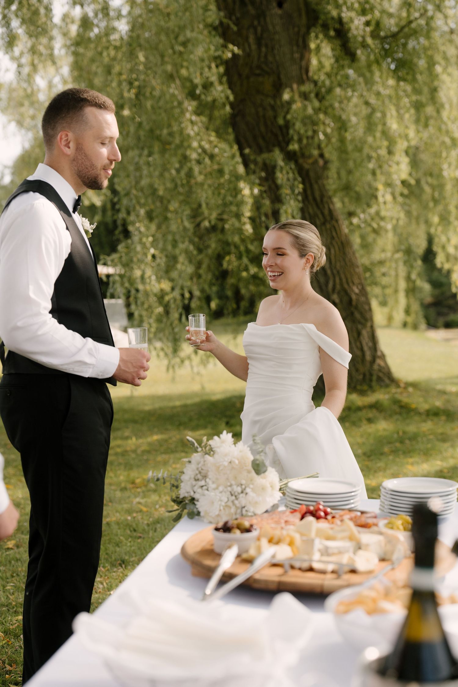 Bride and groom toast with champagne at outdoor wedding reception table under willow tree.