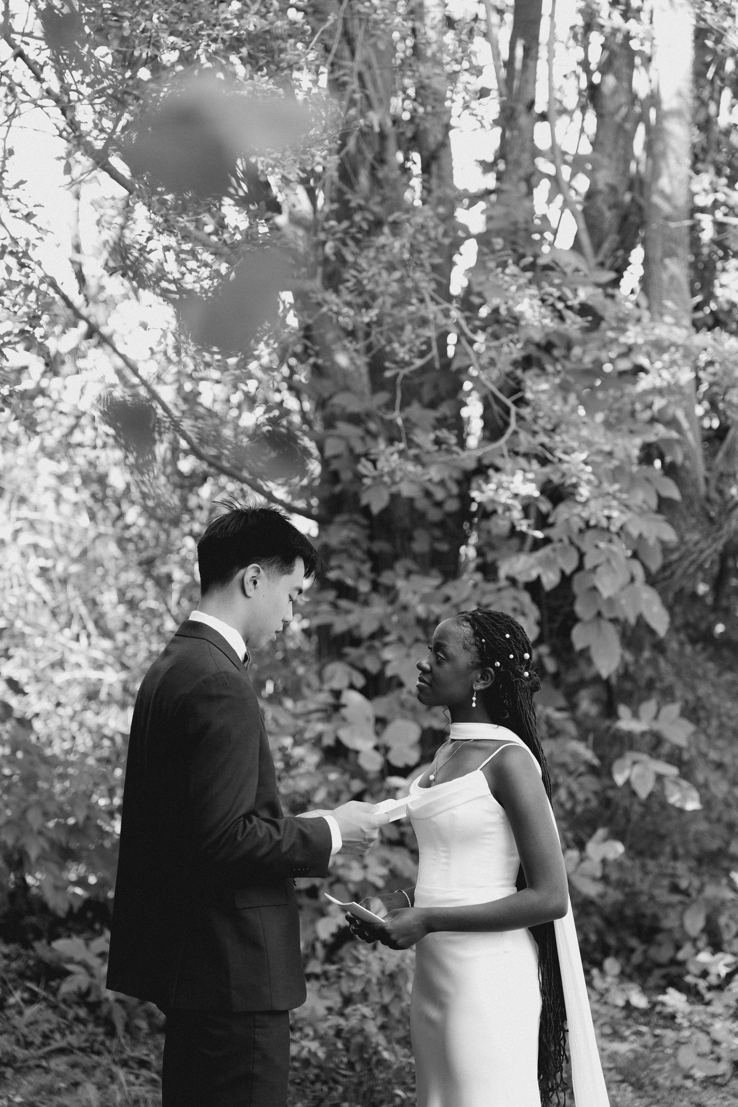 A black and white photo of an interracial couple exchanging wedding vows outdoors surrounded by lush greenery at Wren House.