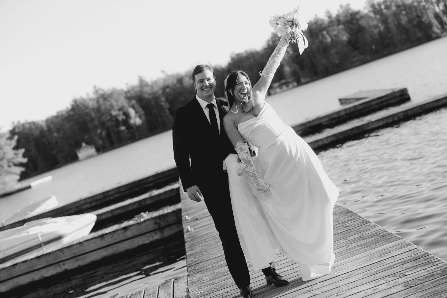 Black and white photo of joyful bride raising bouquet on a dock with groom by a lakeside.