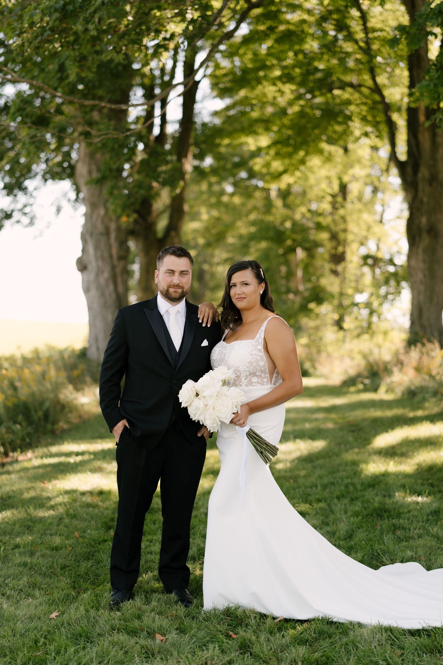 Bride in white mermaid gown holds bouquet beside suited groom outdoors under tall trees.