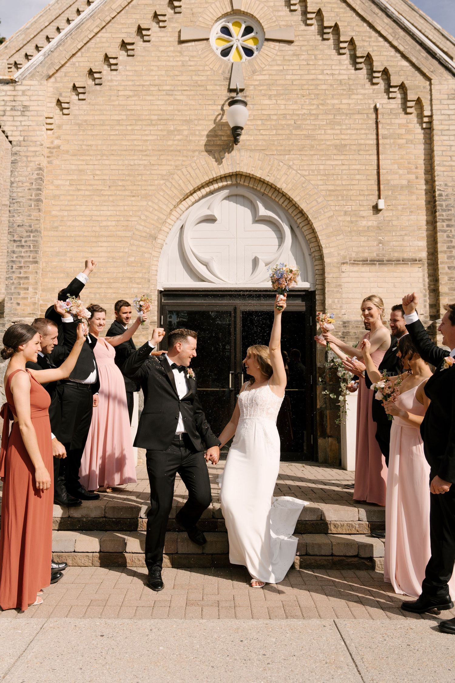 Joyful newlyweds exit a brick church, raising hands in celebration, surrounded by cheering wedding party in pink and black attire.