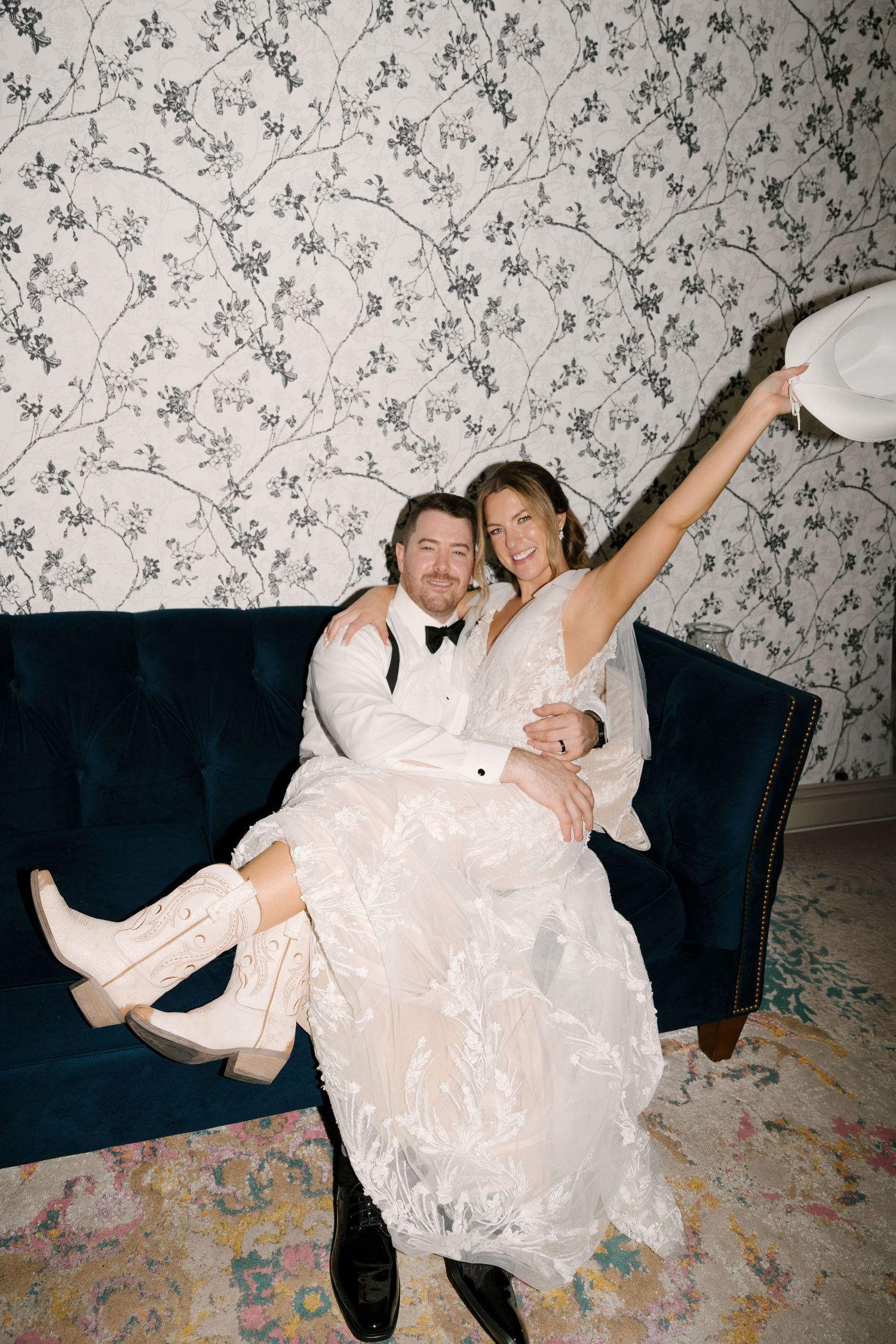 Joyful bride and groom sitting on blue velvet sofa, bride wearing cowboy boots and holding white hat up in celebration inside St Marys Venue