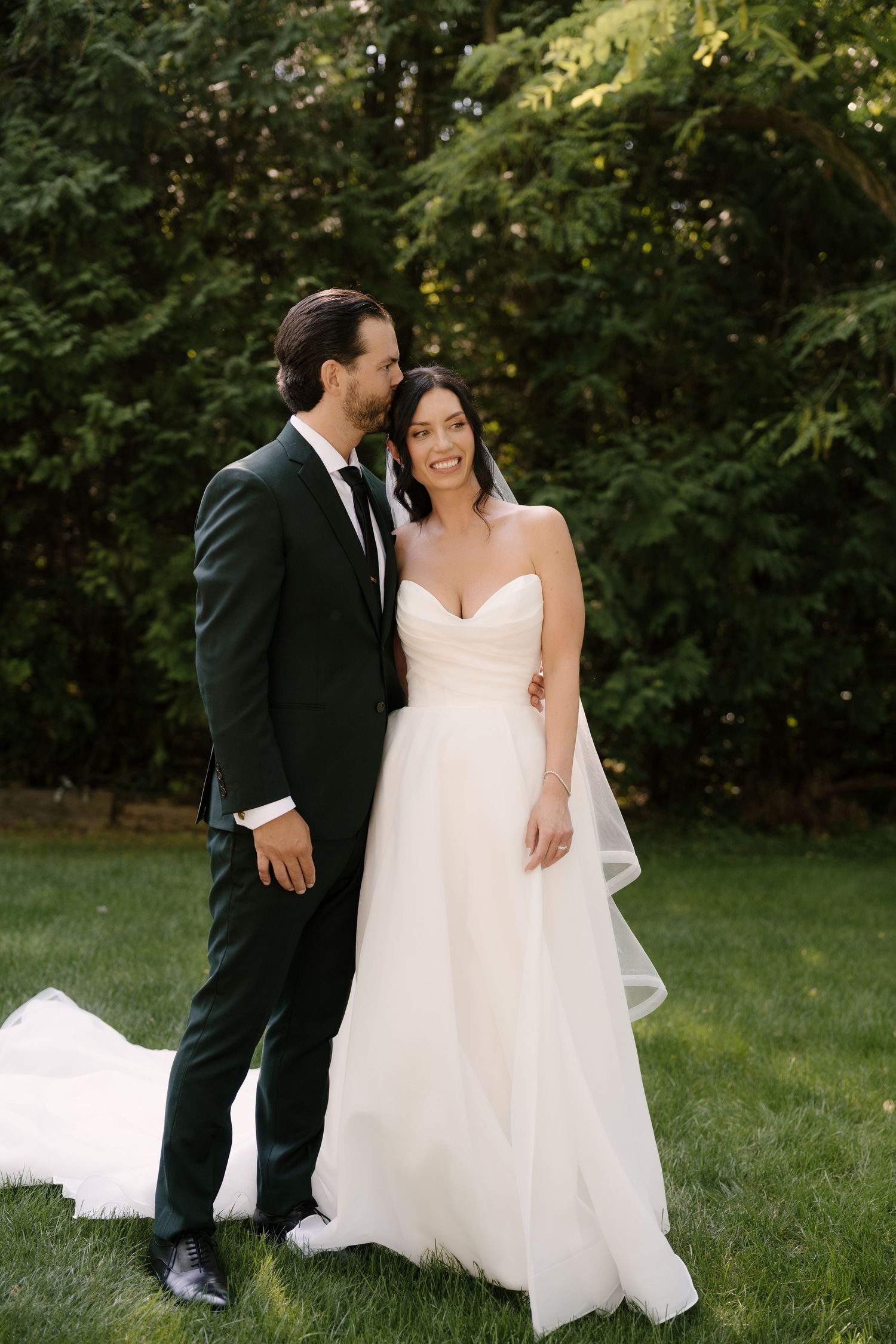 Bride in strapless white gown and groom in dark suit share a romantic moment on green lawn with trees behind.