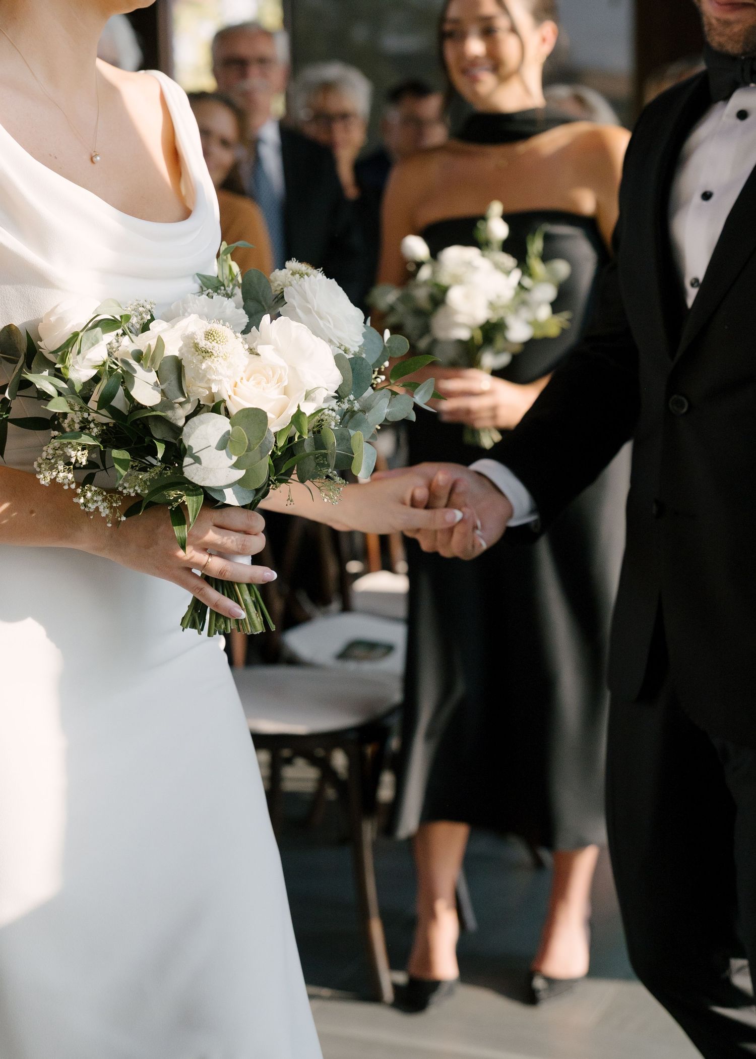 Bride in white cowl-neck gown holding white floral bouquet with greenery, walking with groom in black tuxedo.