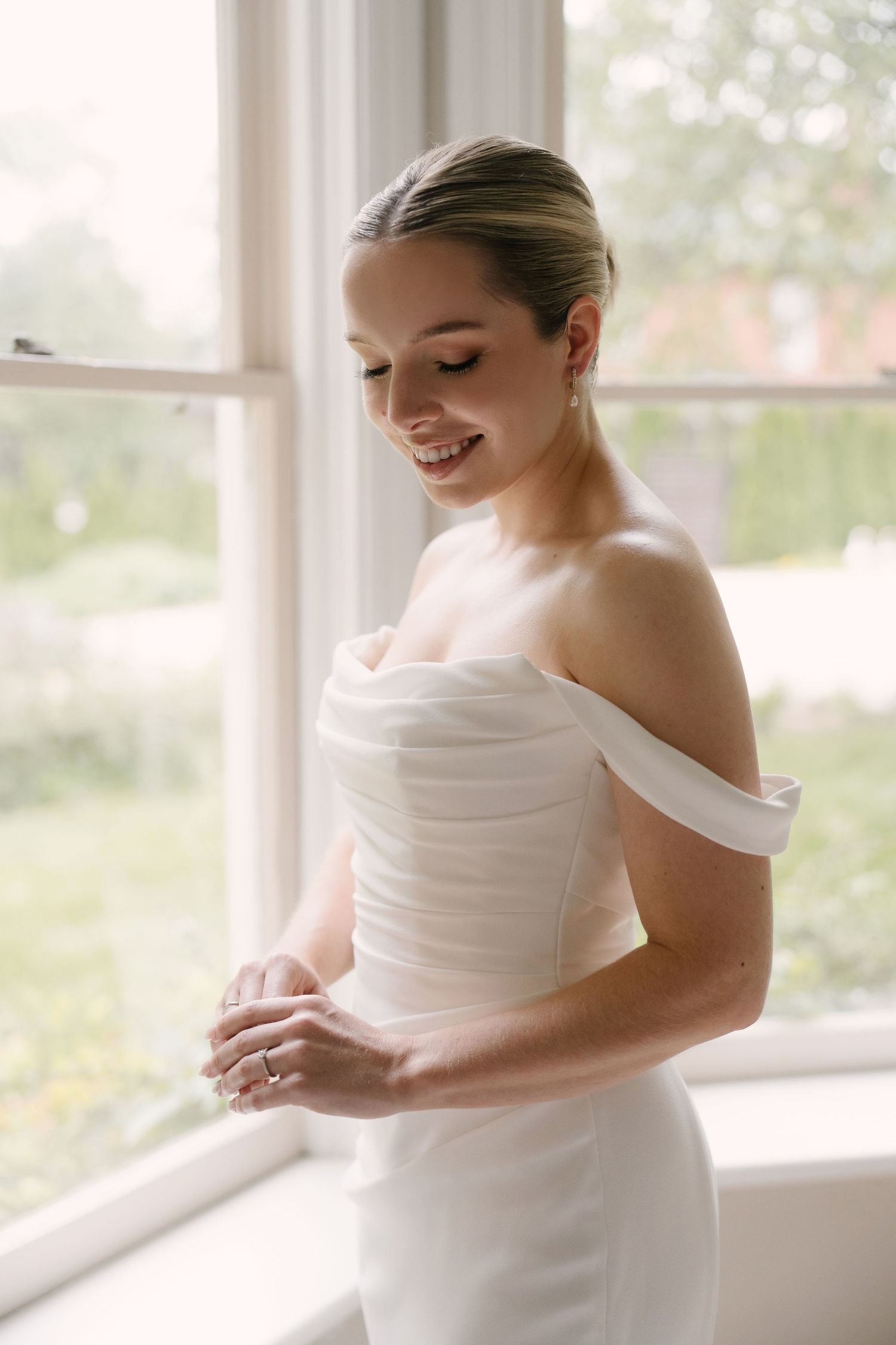 Smiling bride in off-shoulder white wedding gown looking down by a bright window.