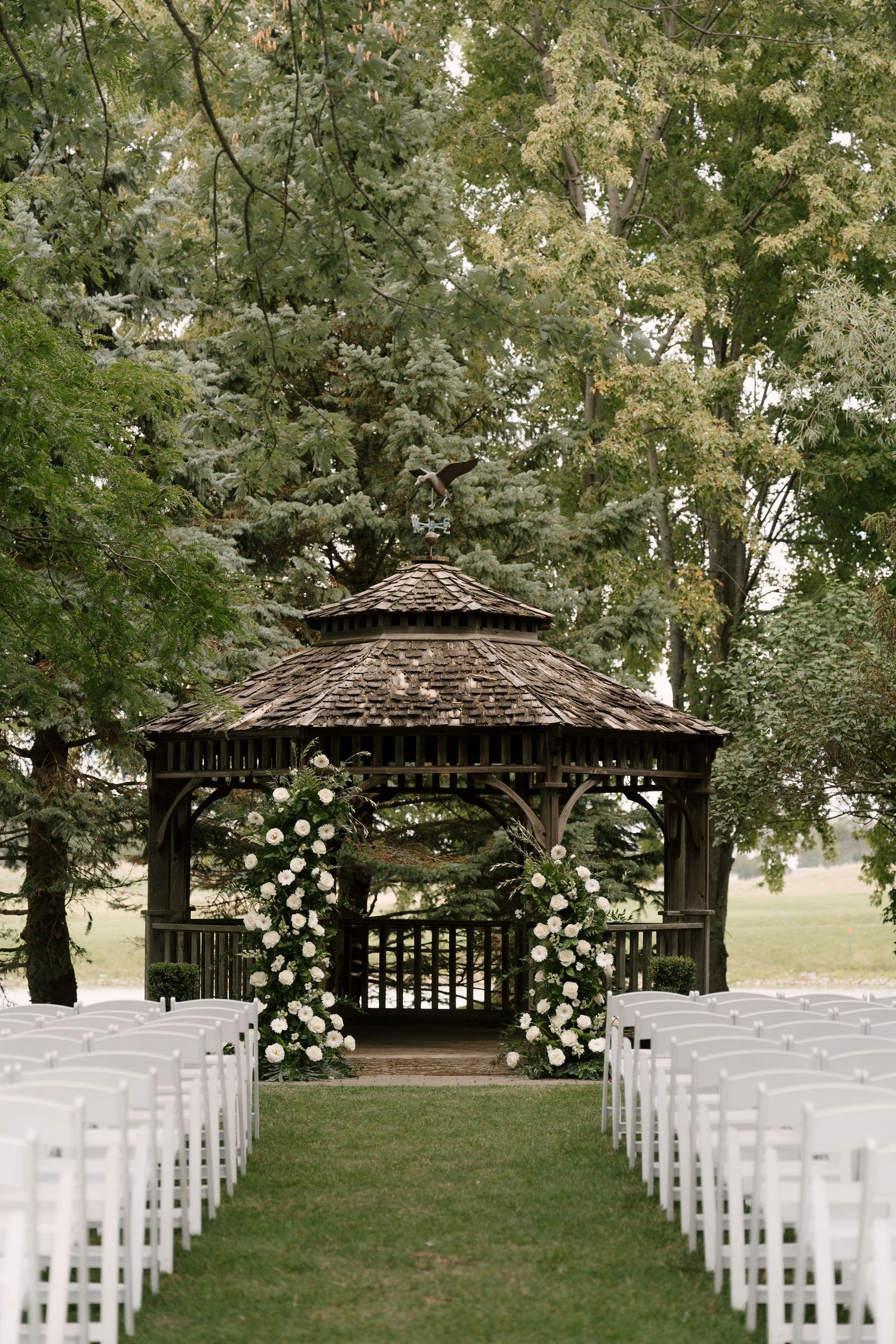 Rustic wooden gazebo adorned with white floral arches flanked by white chairs for an outdoor wedding ceremony.