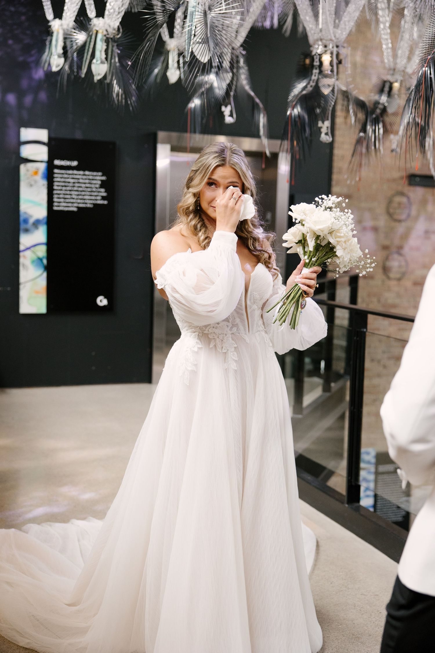 Emotional bride in off-shoulder white tulle gown wiping tears while holding a white floral bouquet indoors.