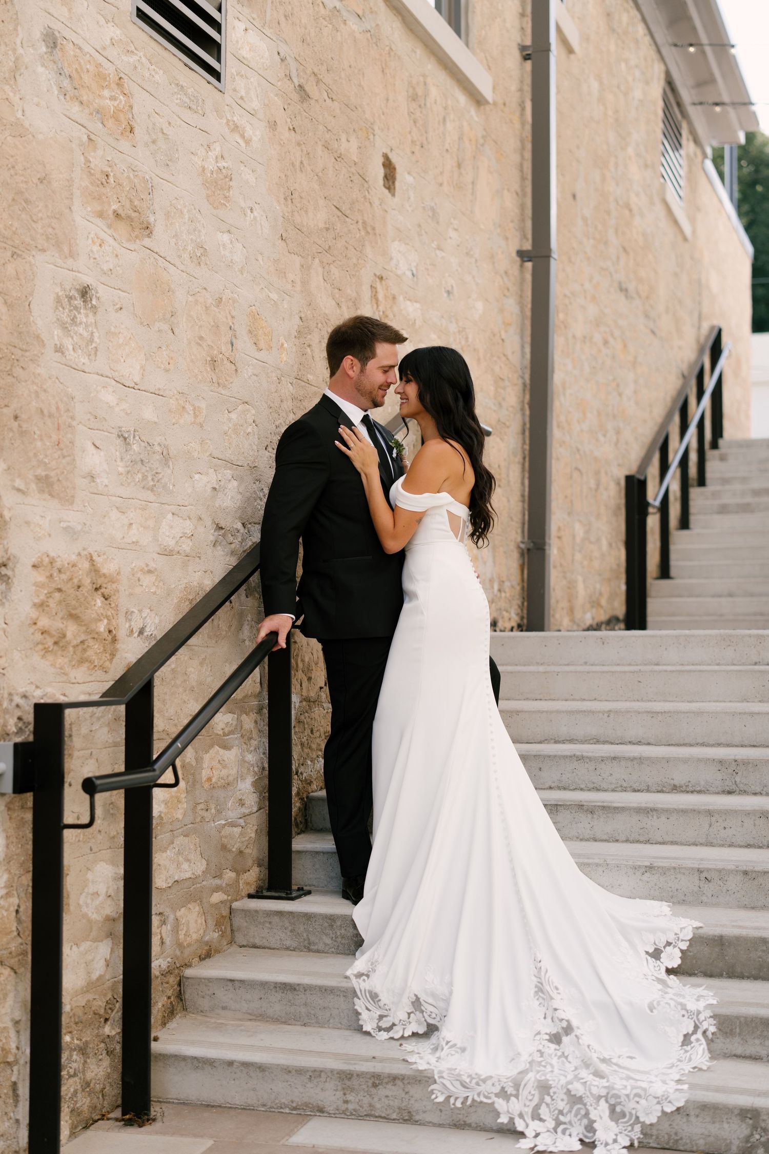 Bride and groom share a romantic moment on stone steps, bride in off-shoulder lace train gown, groom in black tuxedo.