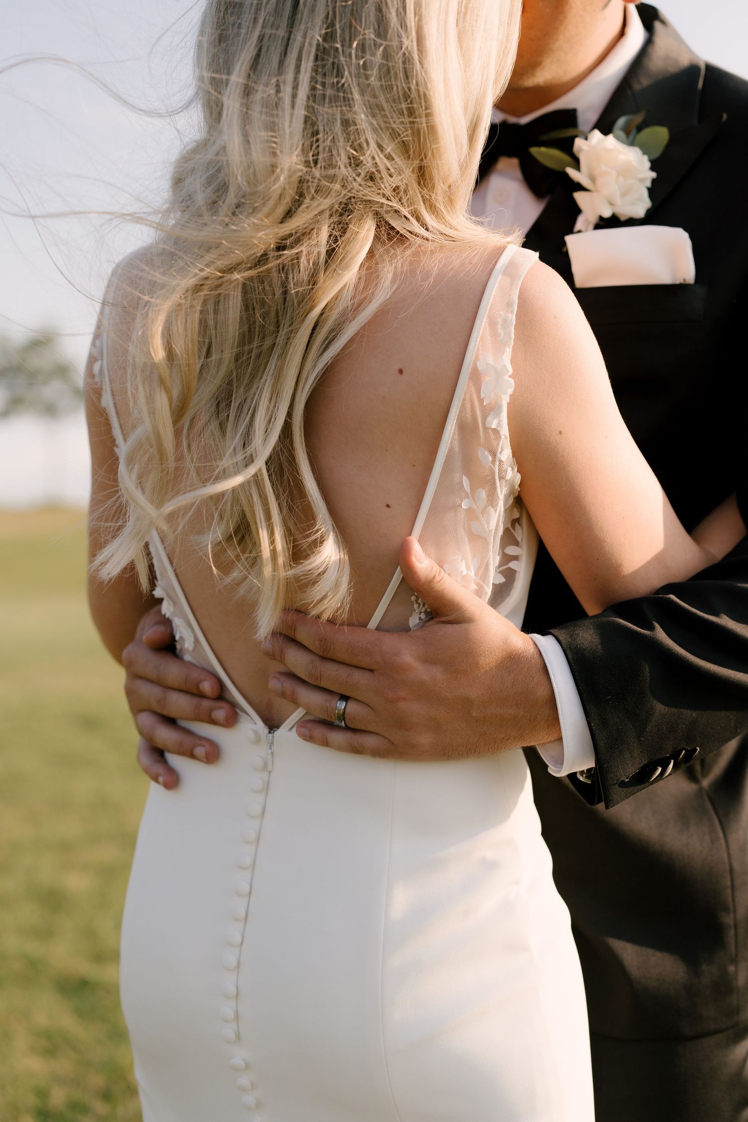 Close-up of bride's backless lace wedding gown with button detail as groom holds her waist.