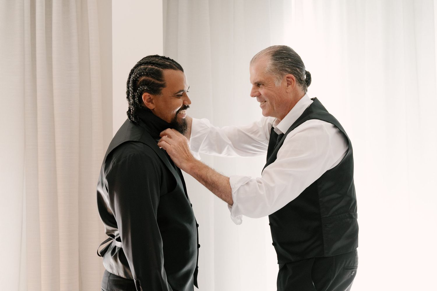 Older man in vest adjusts groom's collar as he prepares in black suit before wedding ceremony at The Walper Hotel.