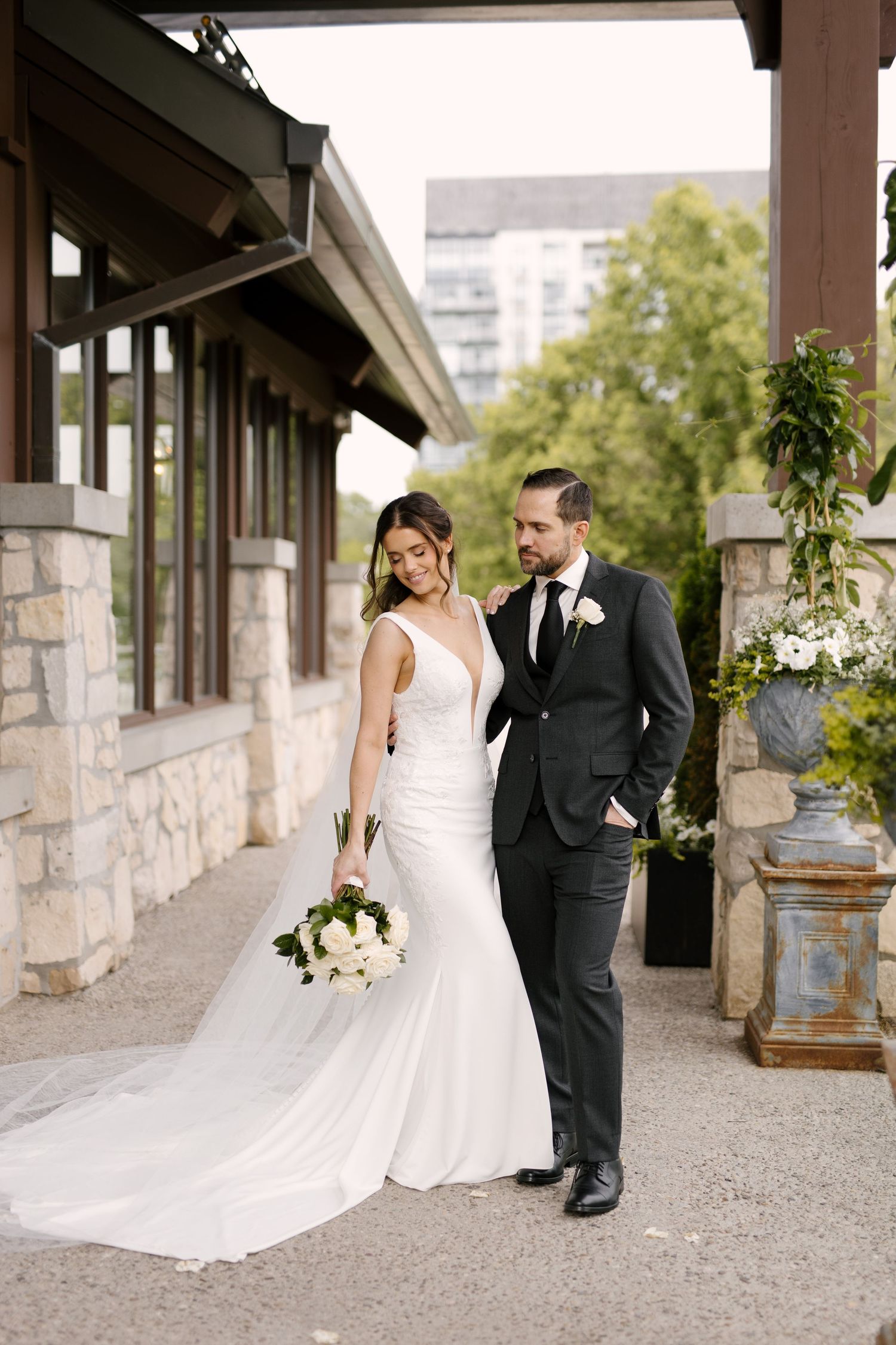 Bride in white mermaid gown holding bouquet walks with groom in black suit along stone Cambridge Mill venue exterior.