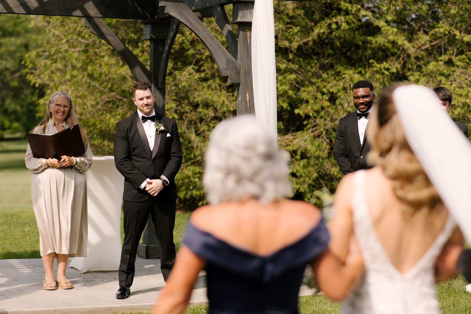 Groom watching bride walk down the aisle during outdoor wedding ceremony under wooden pergola at St Marys Wedding Venue.