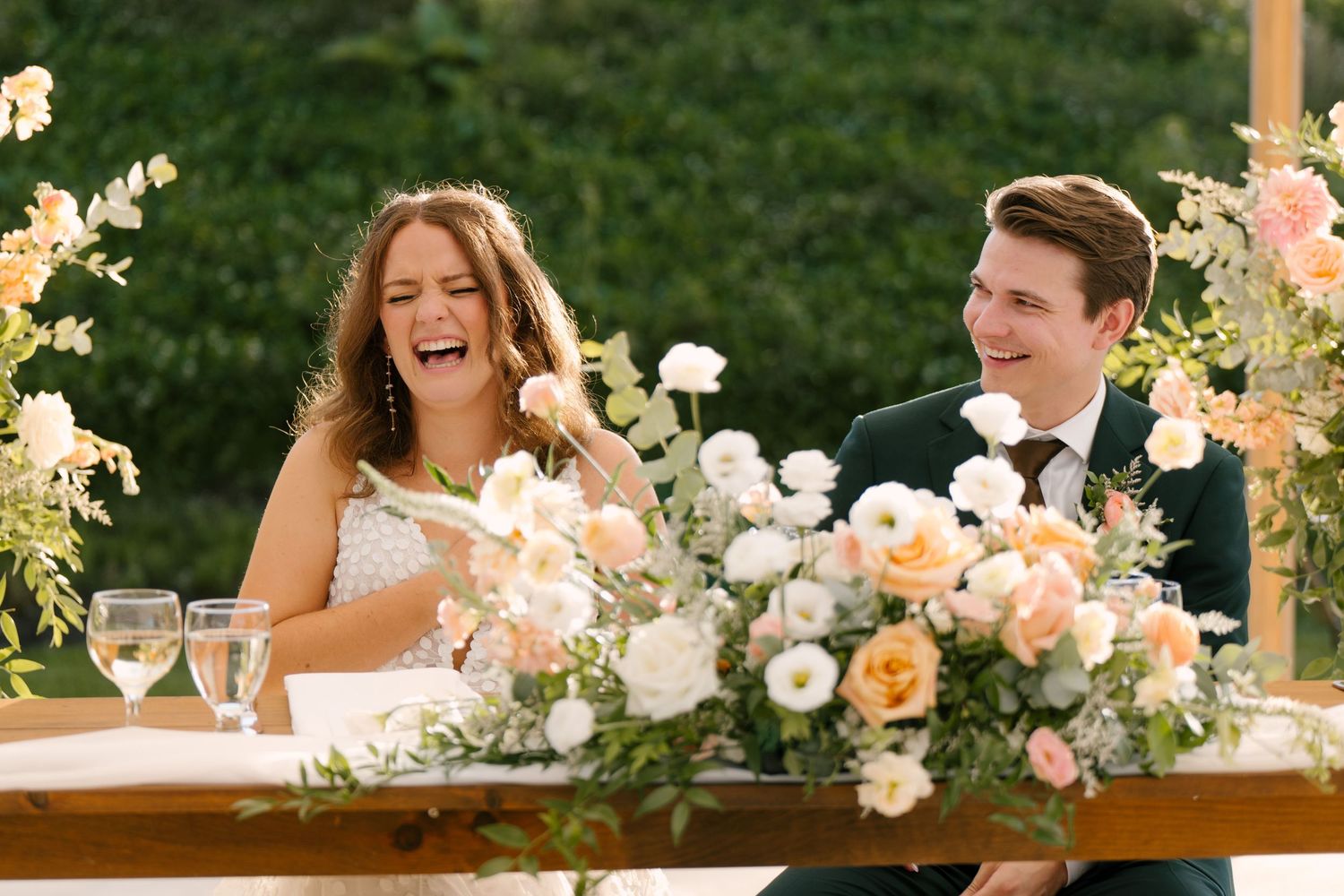 Laughing bride and groom seated at sweetheart table adorned with peach and white floral arrangements. Under Wakefield Estates tent reception