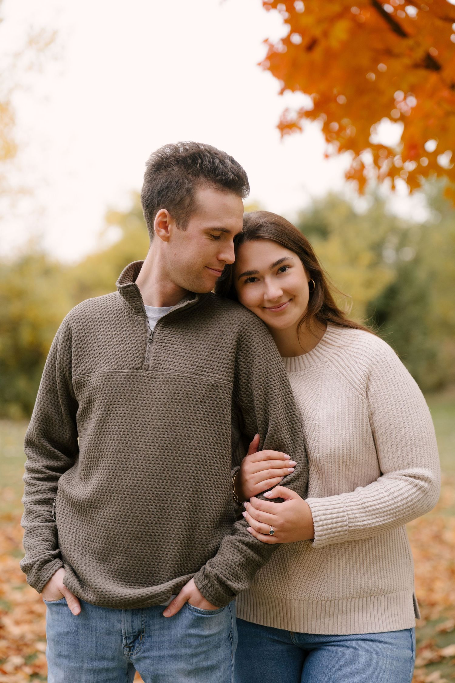 Couple embracing in autumn park, woman smiling at camera, colorful fall foliage in background.