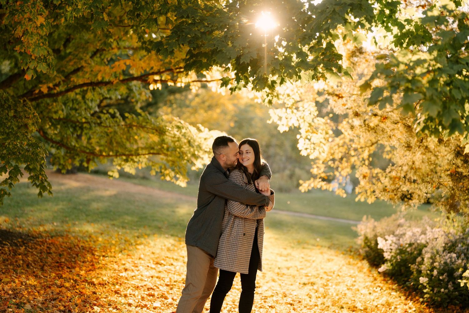 Romantic couple hugging on a leaf-covered path in a glowing autumn park at golden hour.