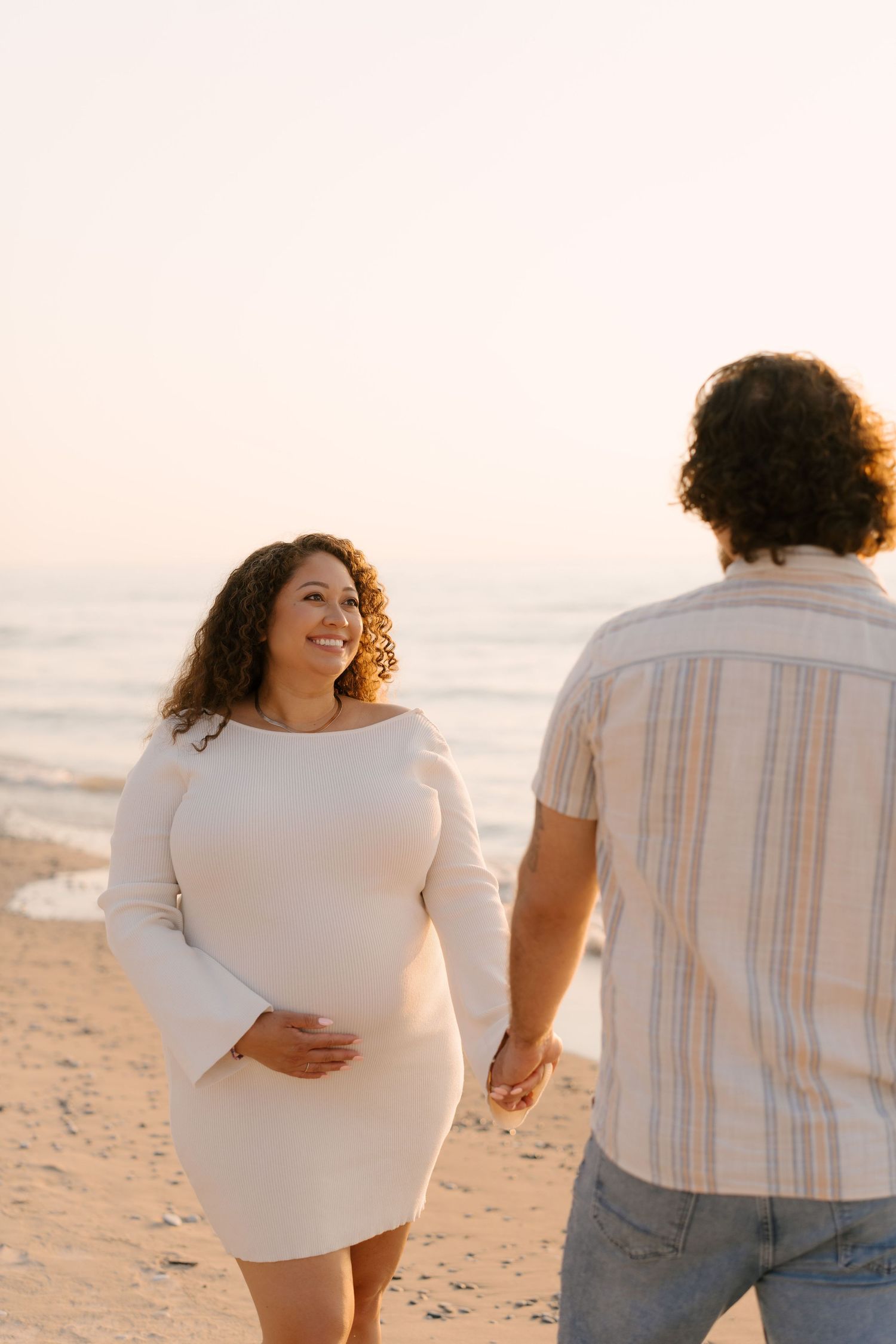 Glowing pregnant woman in white dress holds bump while walking with partner on beach.