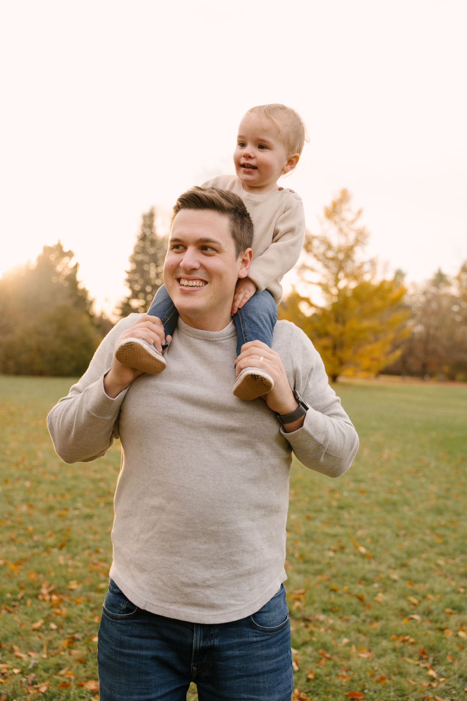 Smiling father in gray sweater carrying happy toddler on shoulders in fall park.