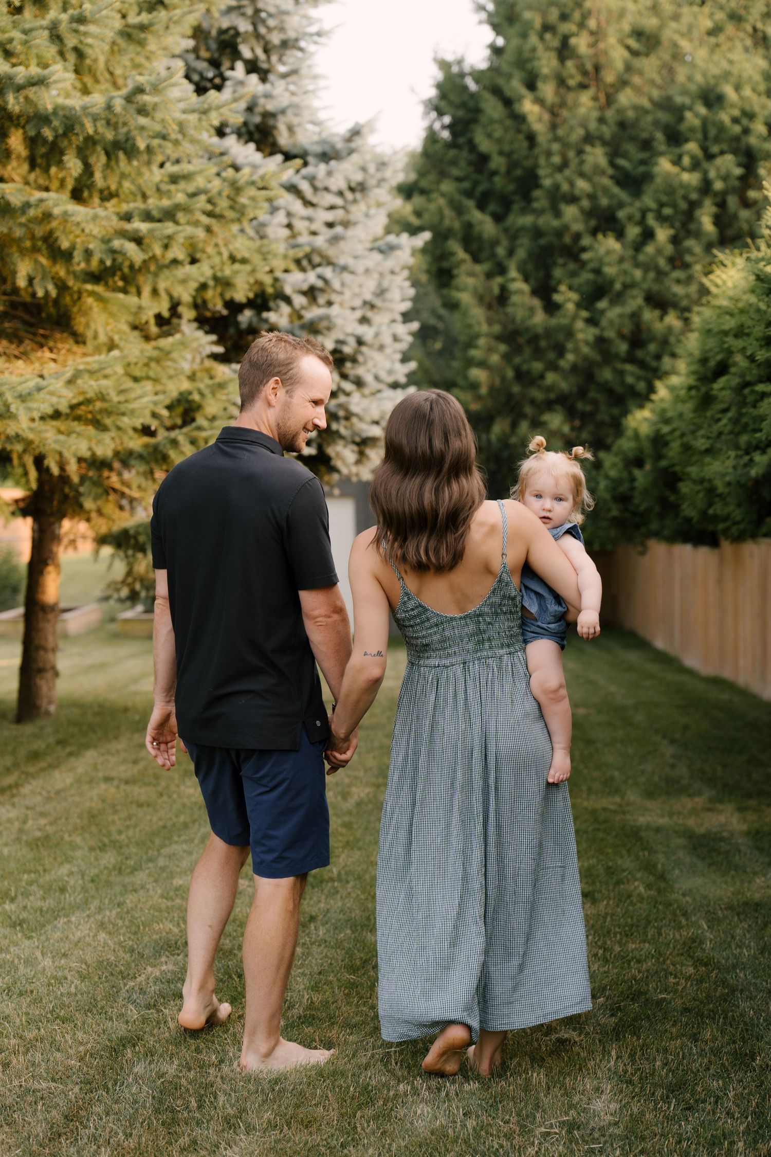 A family walks hand in hand through a lush green yard, mother carrying their toddler daughter.