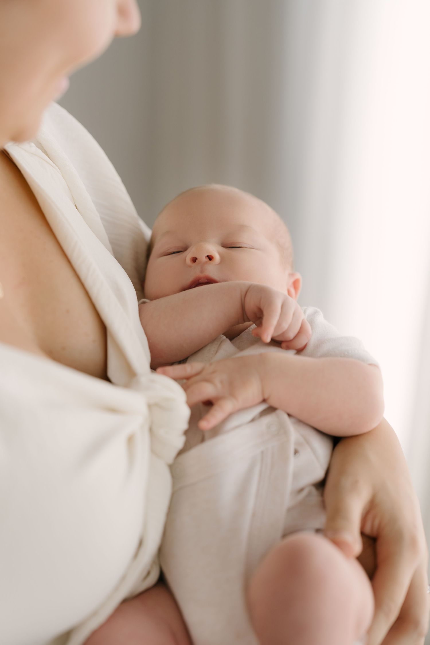 Mother in white dress gently holding sleeping newborn baby against her chest in soft light.