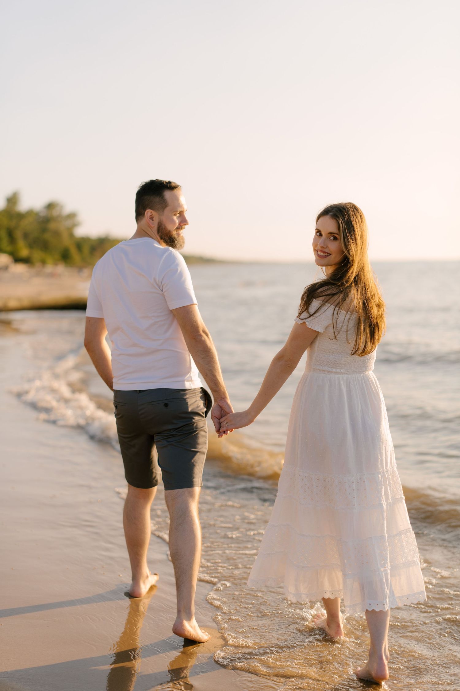 Couple holding hands on a sandy beach at sunset, woman in white lace dress smiling over her shoulder.