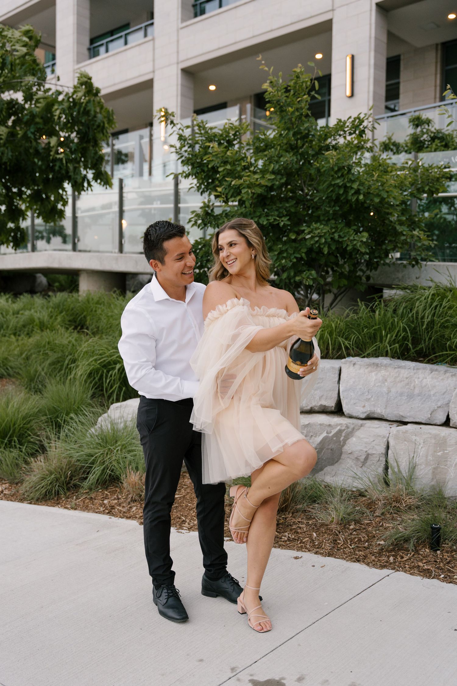 Joyful couple popping a champagne bottle outdoors near a modern building surrounded by lush greenery.