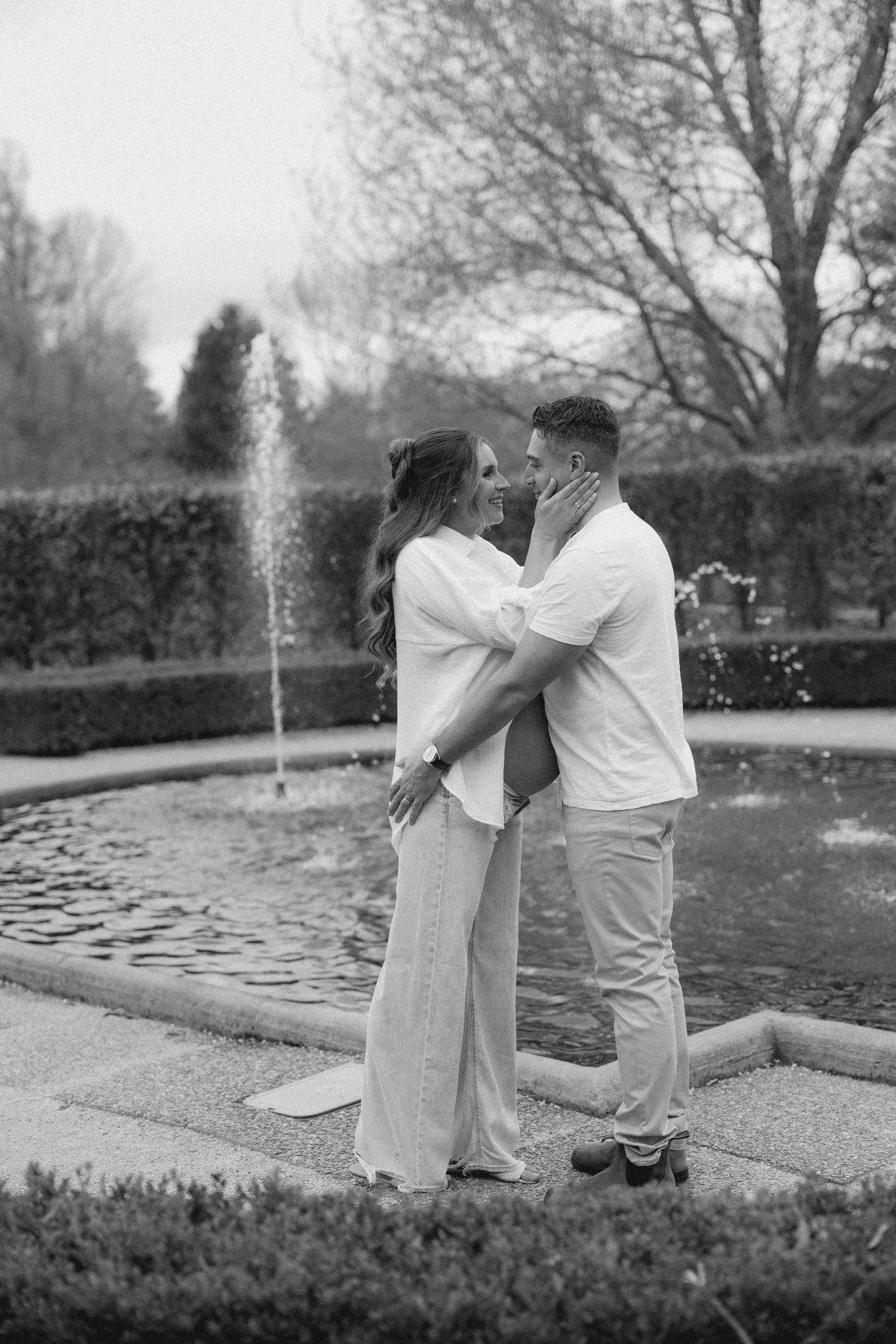 Black and white image of couple embracing lovingly by garden fountain.