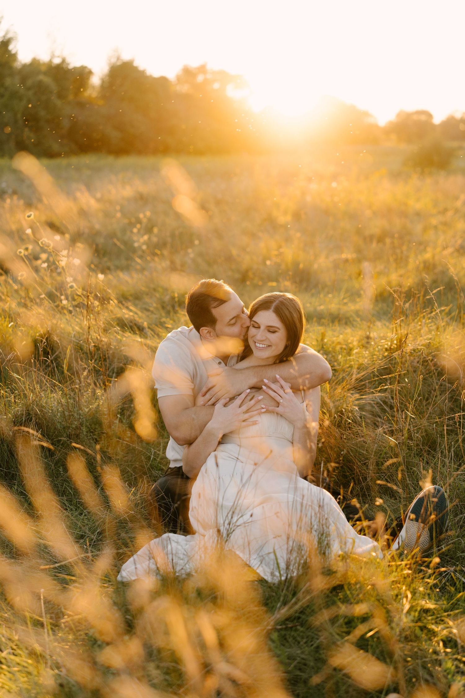Couple sitting together in tall golden grass, embracing warmly during a stunning sunset.
