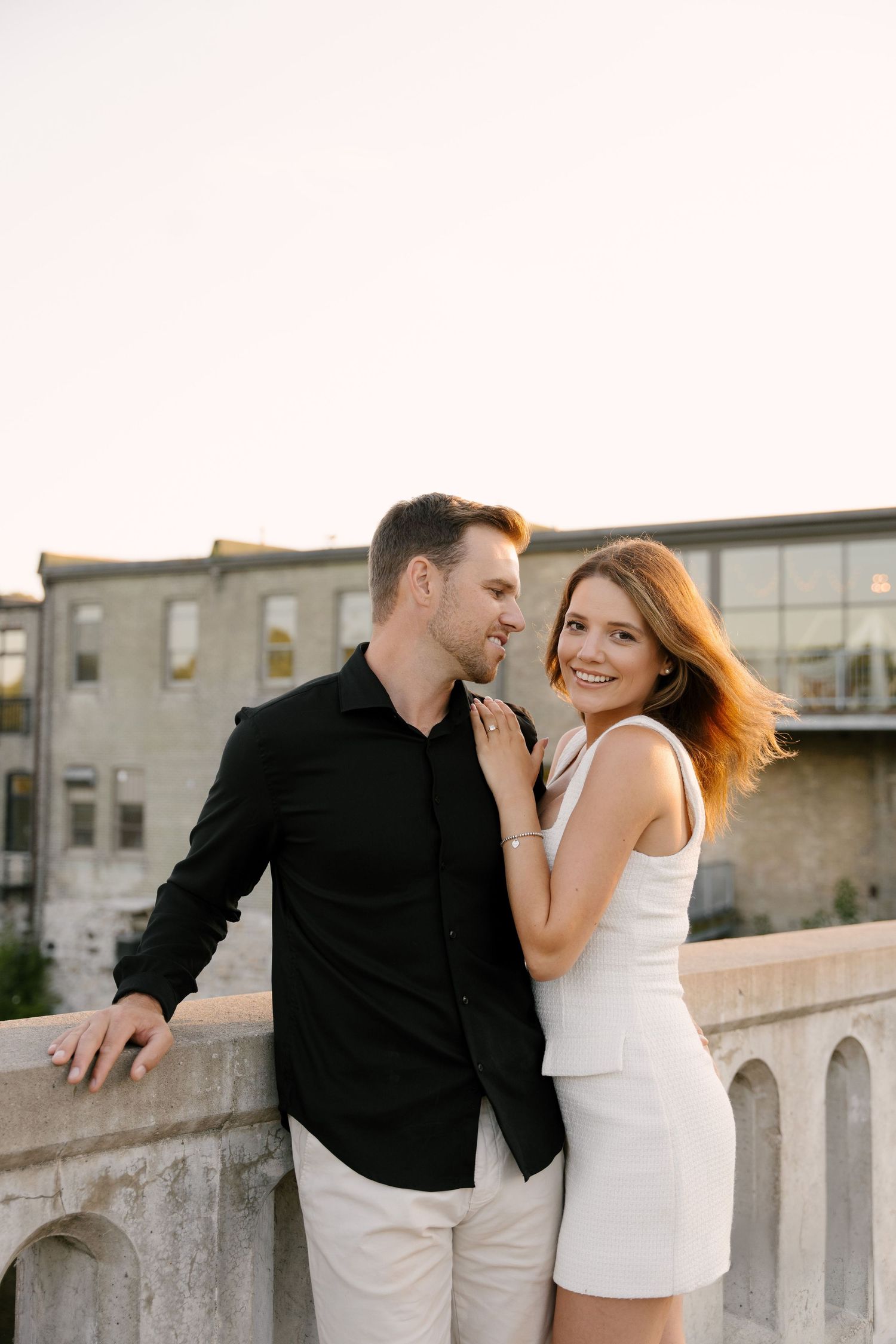 Smiling engaged couple embracing on stone bridge at golden hour sunset.