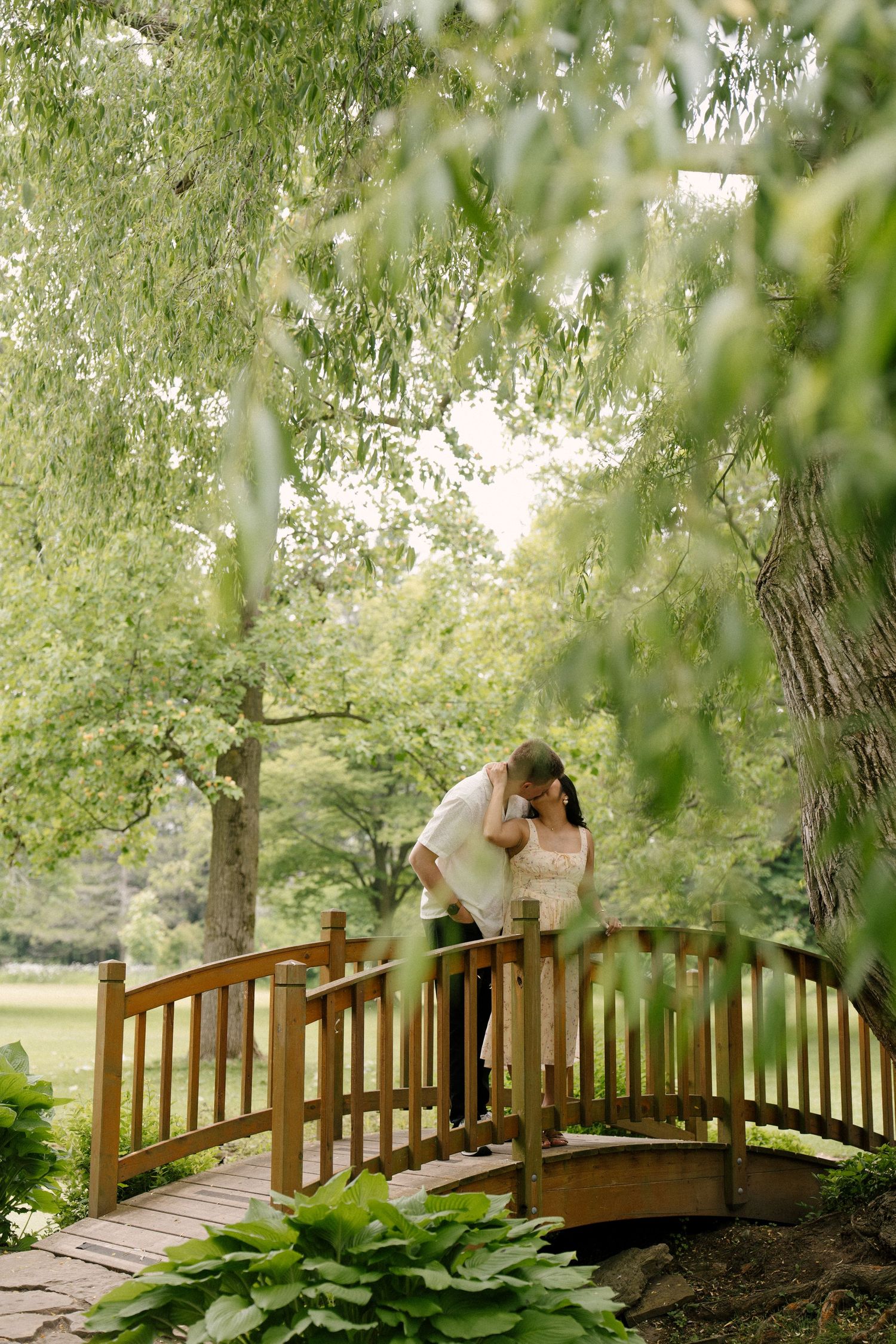 Couple sharing a tender kiss on a wooden garden bridge beneath a large weeping willow tree.