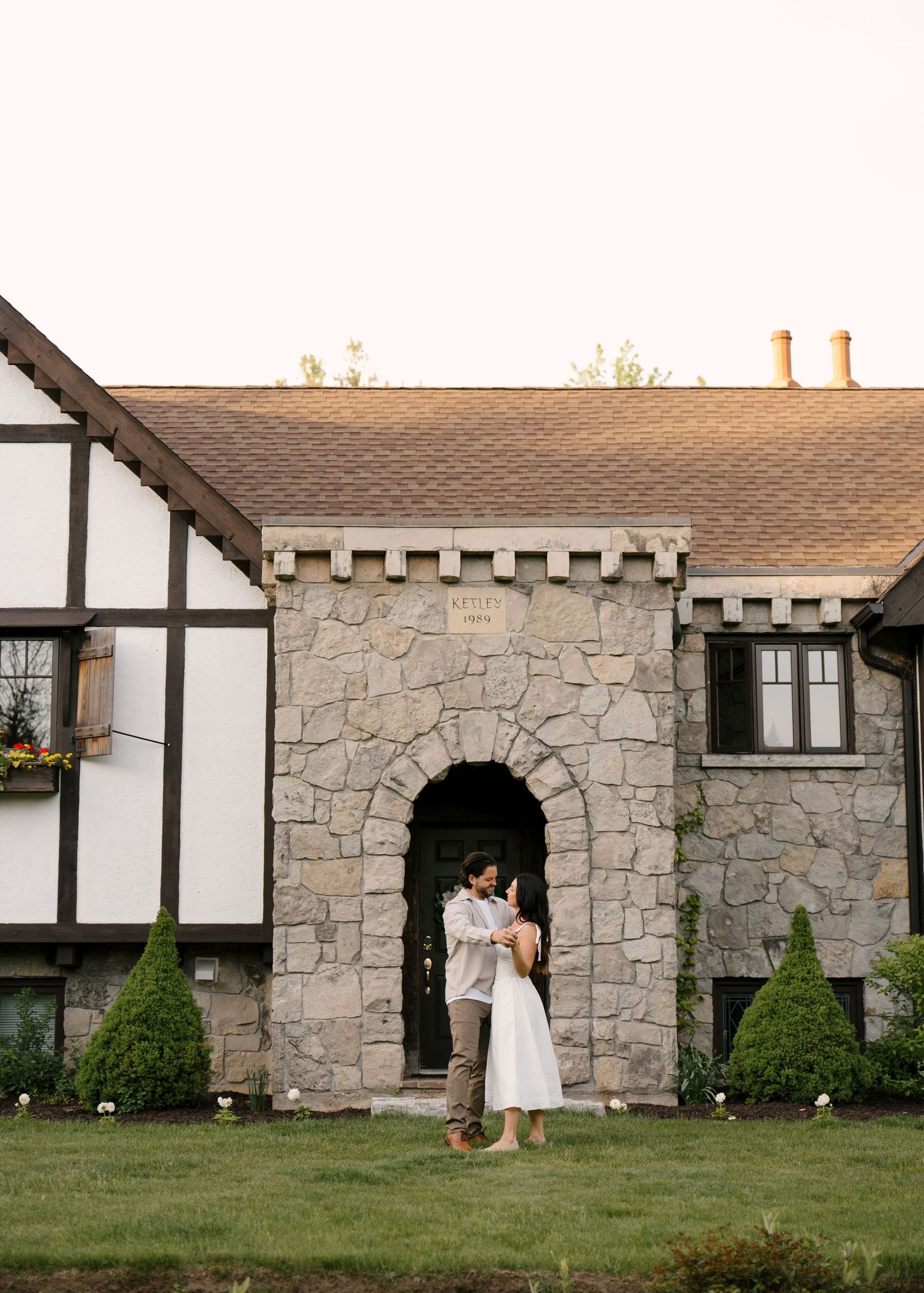 Newlyweds embracing on lawn in front of charming Tudor-style stone building with arch entry.