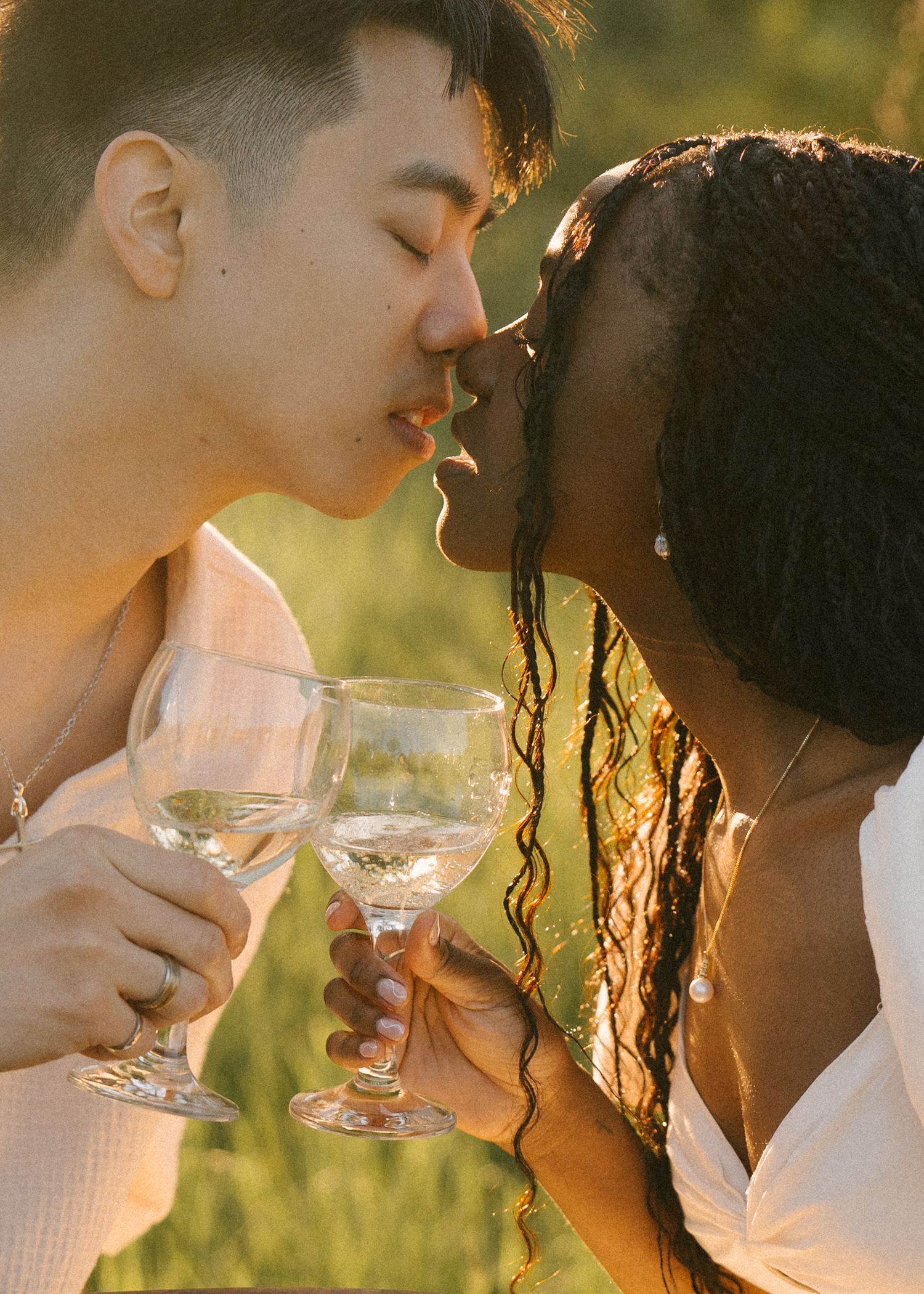 Interracial couple kissing while holding wine glasses in warm golden sunlight.