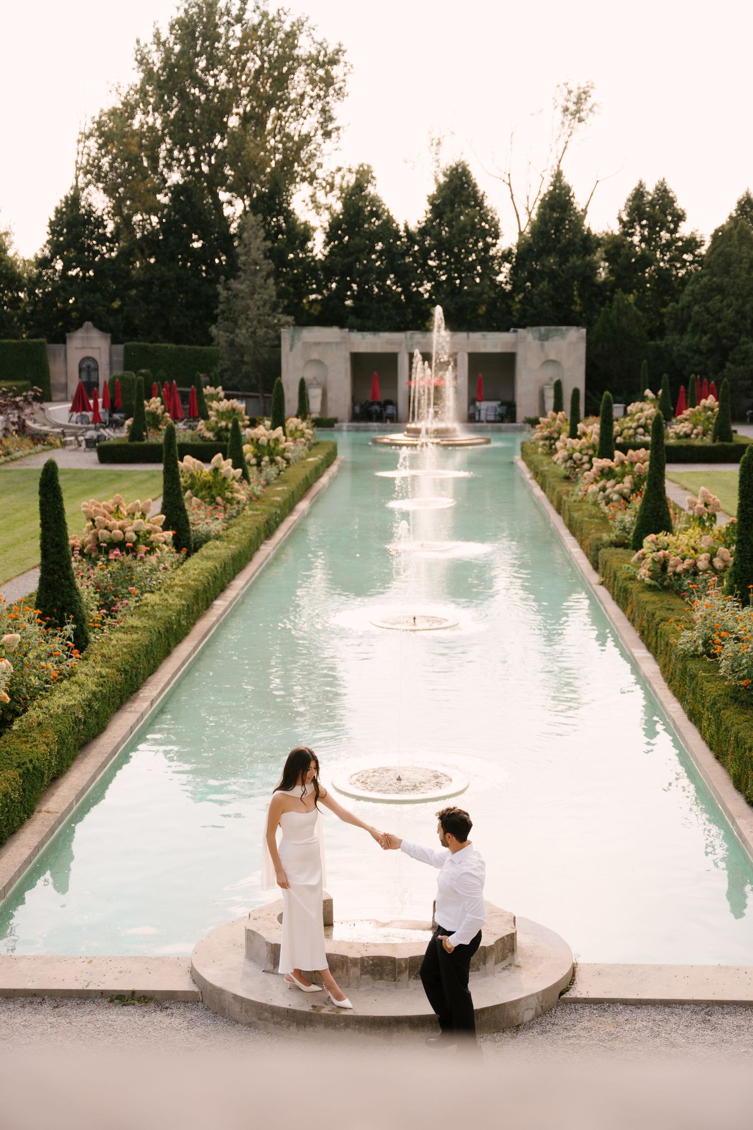 Couple holding hands beside a long formal reflecting pool with fountain in an elegant symmetrical garden at sunset.