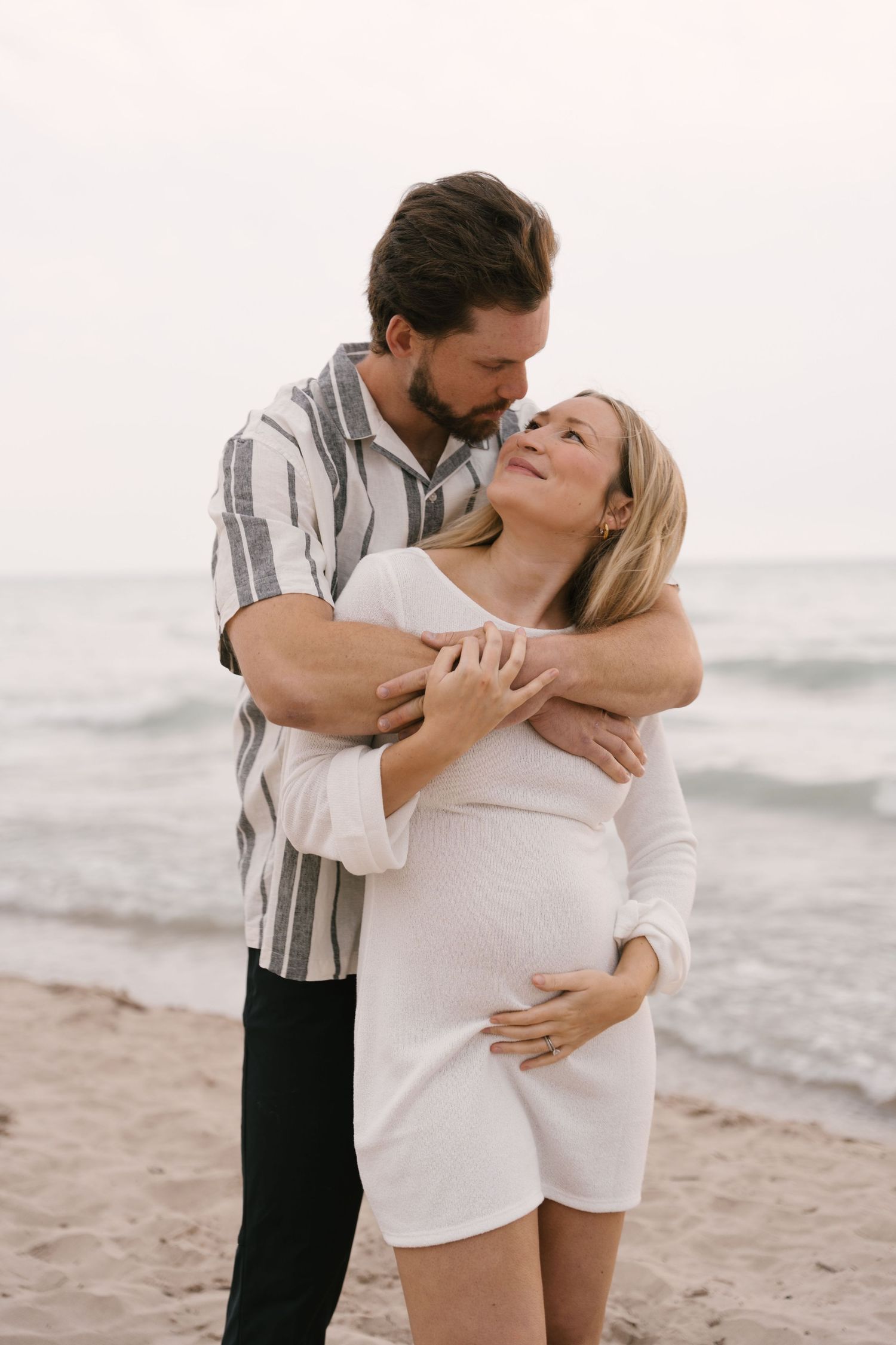 Happy pregnant couple embracing and smiling at each other on a sandy beach.