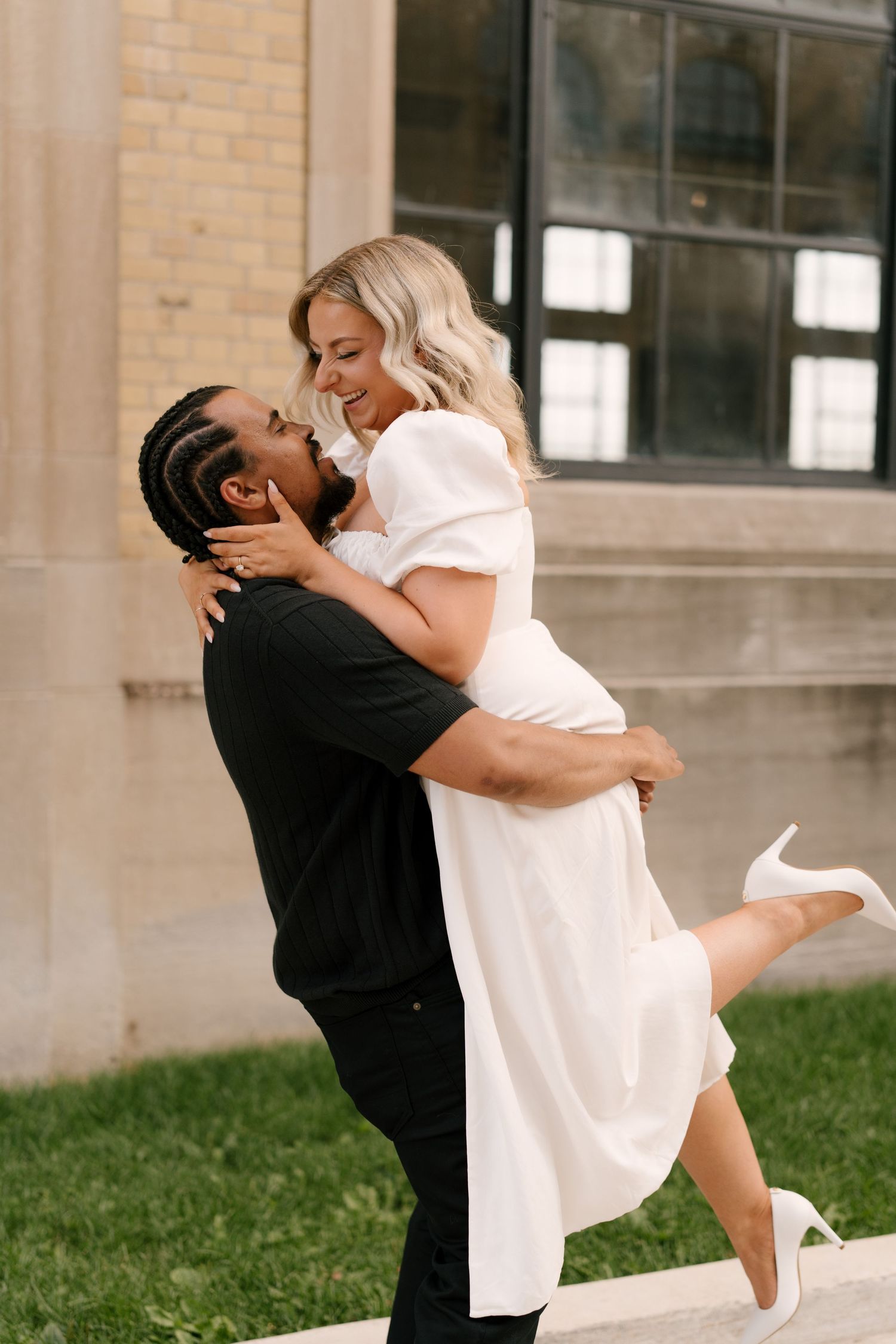 Man in black shirt lifting smiling woman in white midi dress during outdoor engagement photo session.
