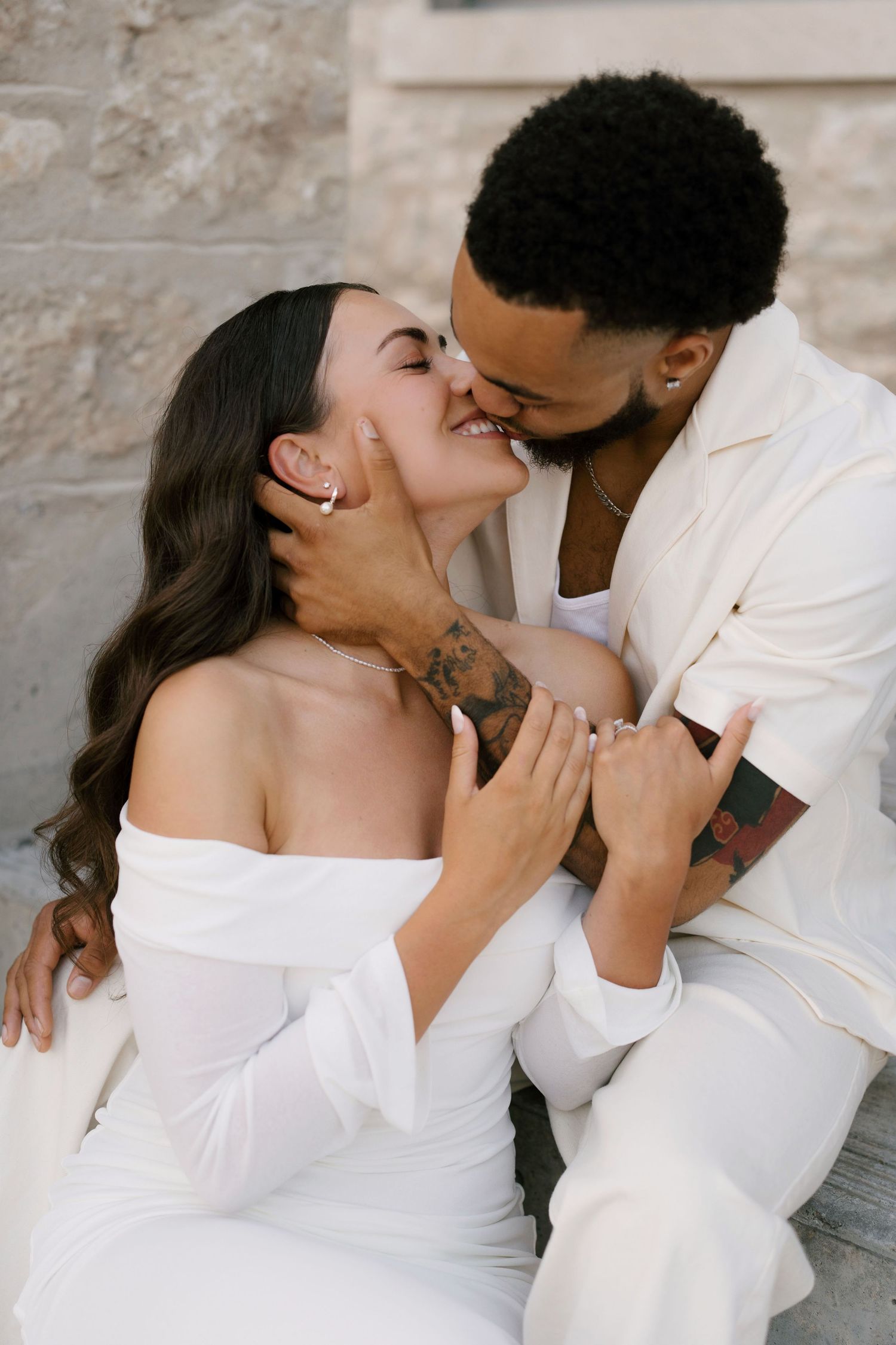 Romantic couple nearly kissing on steps, woman in white off-shoulder dress smiles with eyes closed.