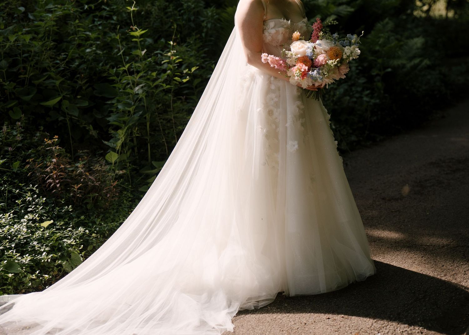 Bride in a floral lace ballgown wedding dress holds a colorful bouquet while standing in a garden setting.