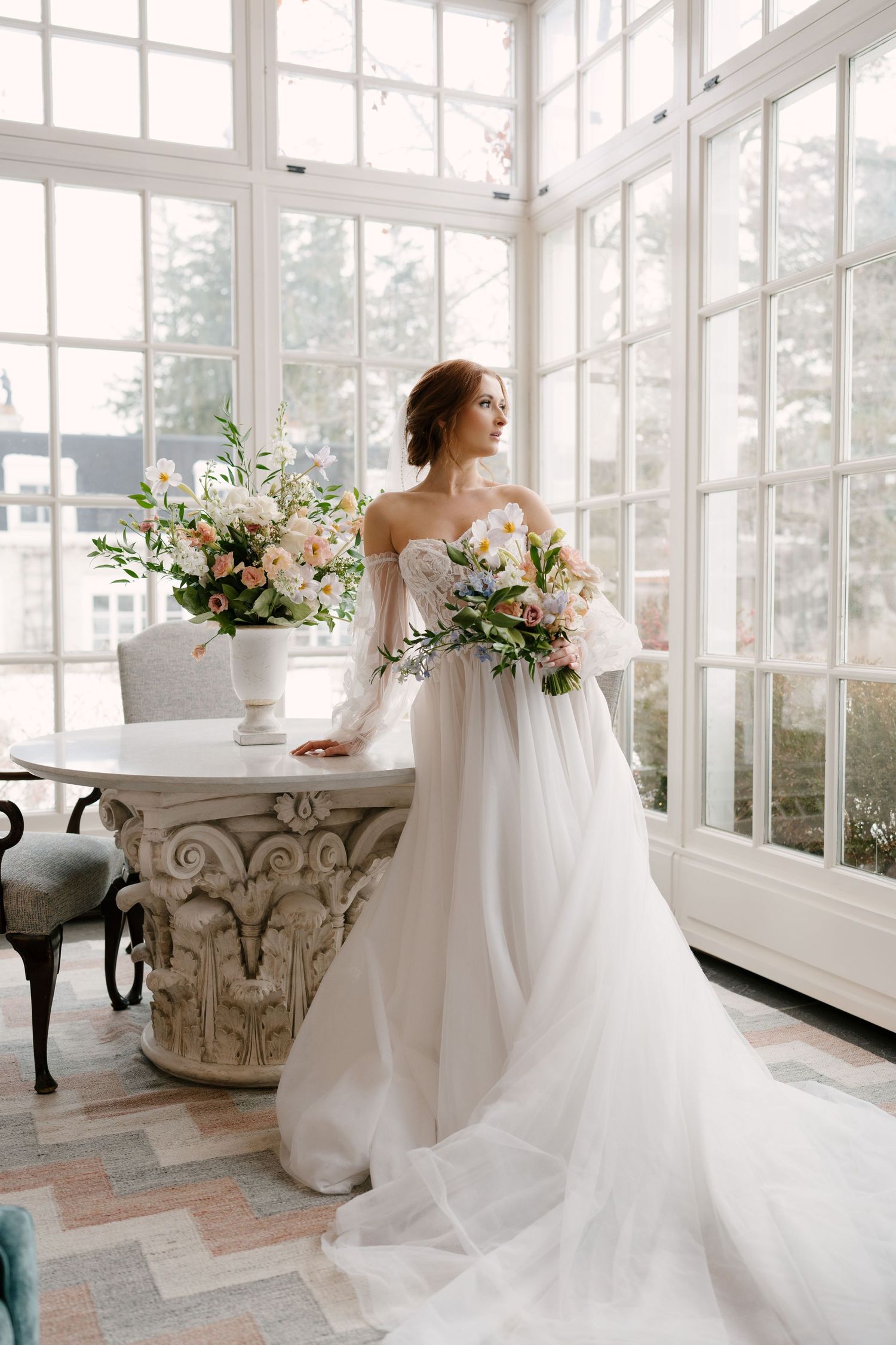 Elegant bride in an off-shoulder gown with a long train stands by large windows holding flowers at Langdon Hall wedding venue.