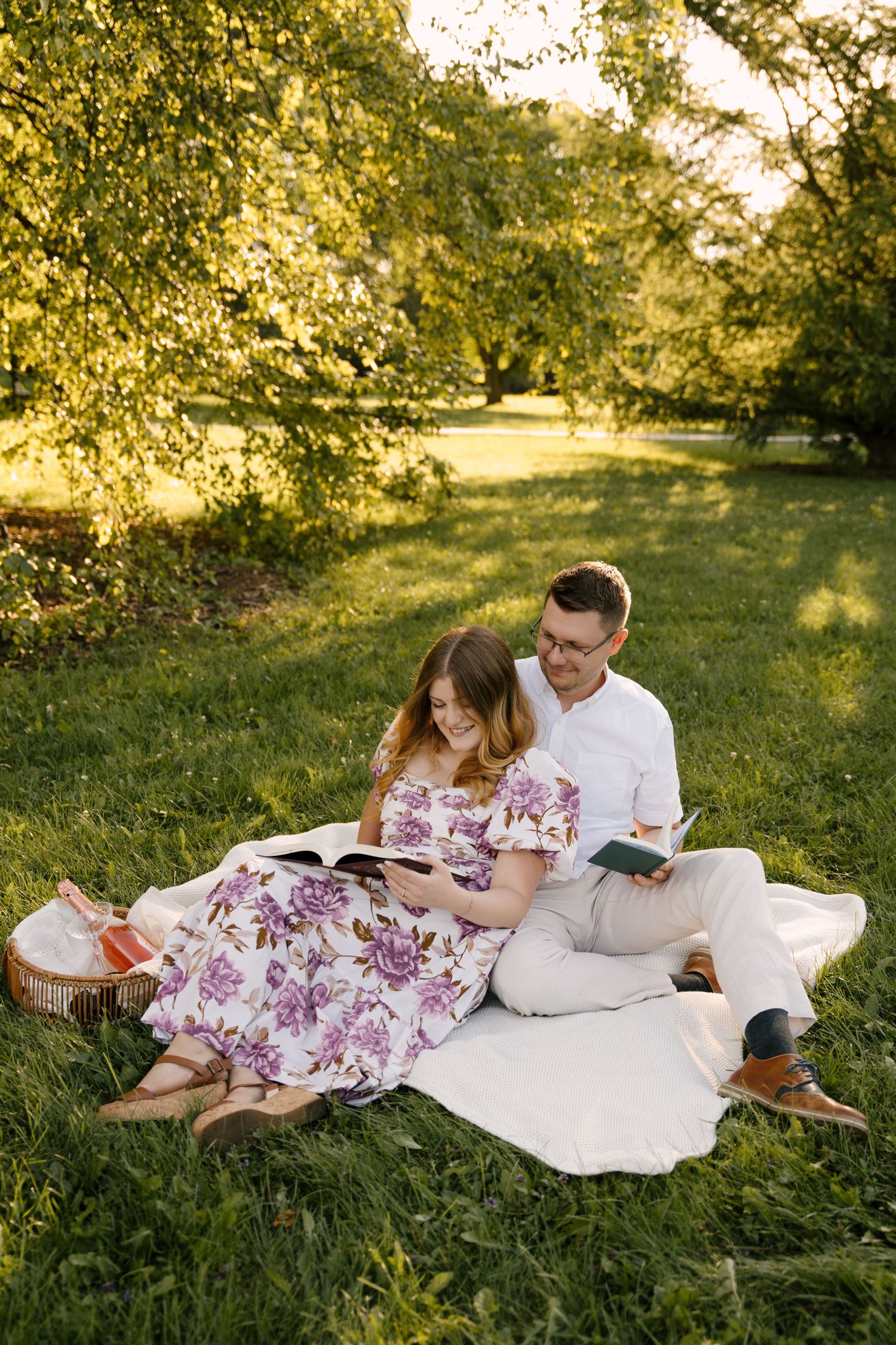 Couple reading books together on a blanket in a sunlit park, woman in a floral dress.