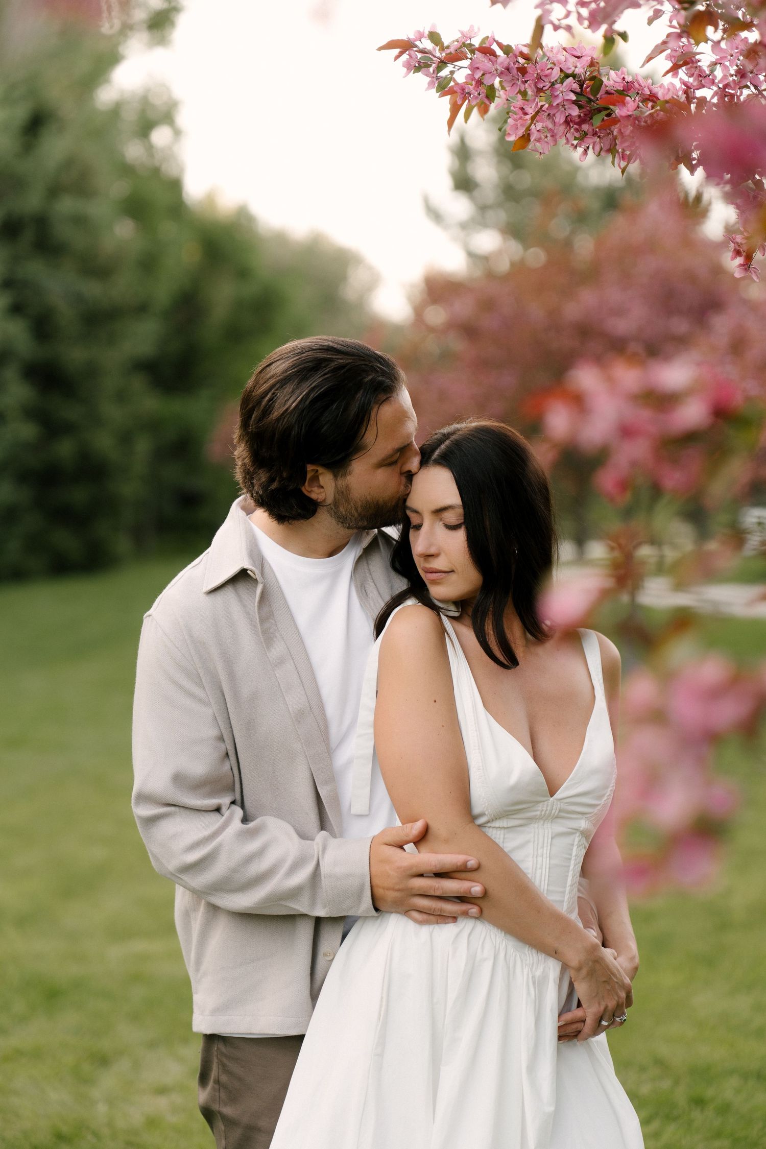 Man tenderly embracing woman from behind near blooming pink trees in a lush green park.