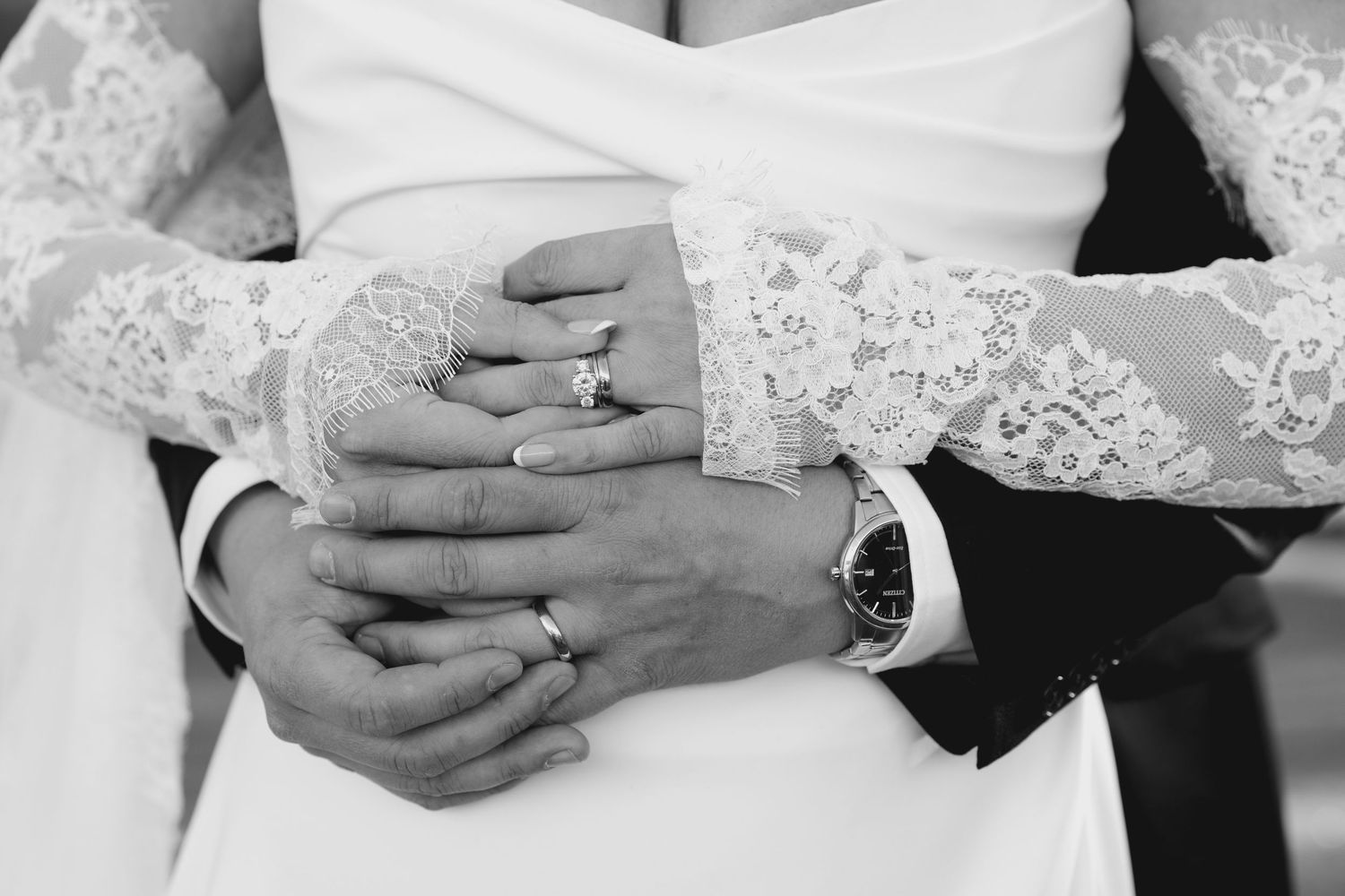 Black and white photo of bride and groom holding hands, showcasing wedding rings and lace bridal gown sleeves.