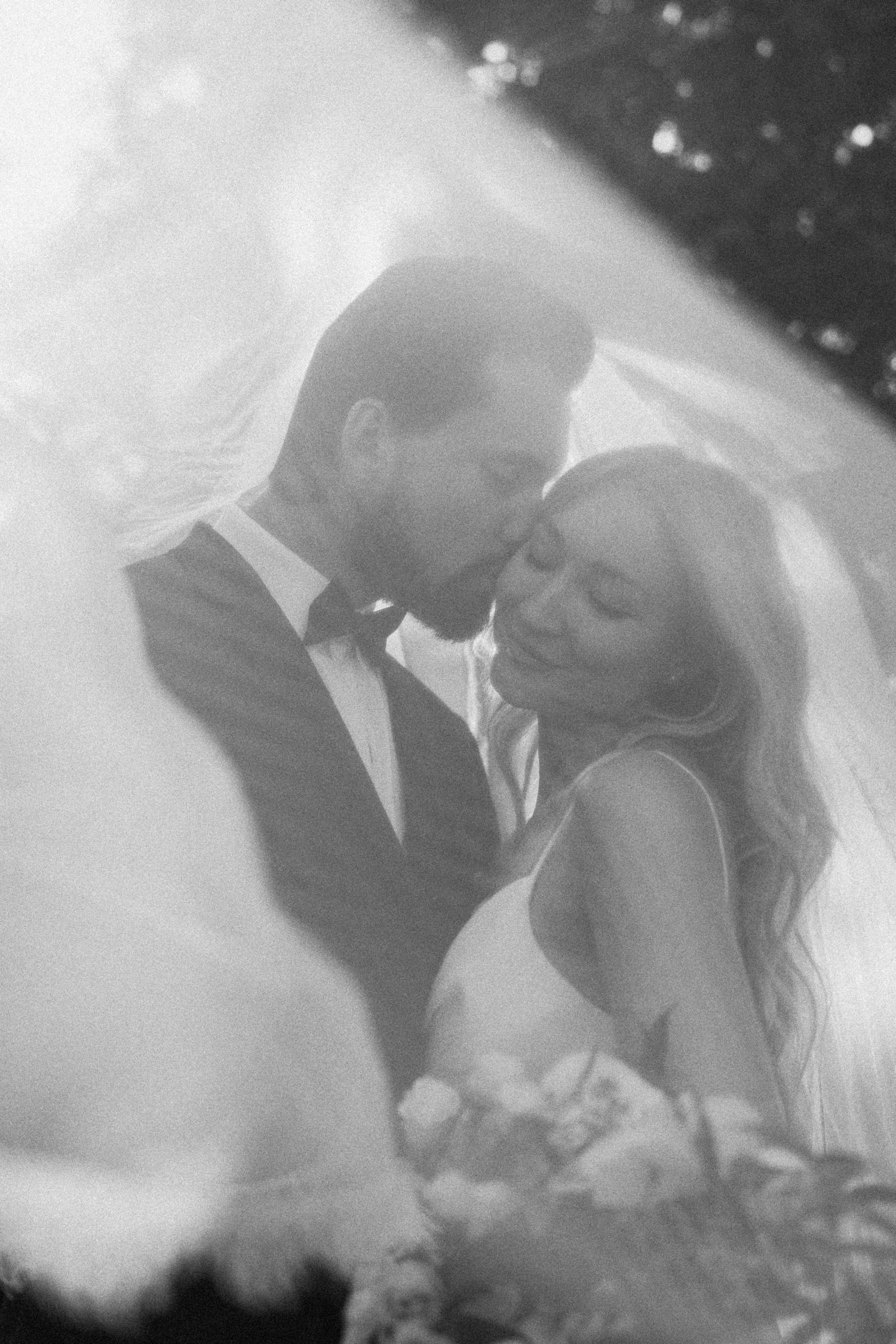 Dreamy black and white wedding portrait of couple almost kissing under a glowing veil in soft natural light.