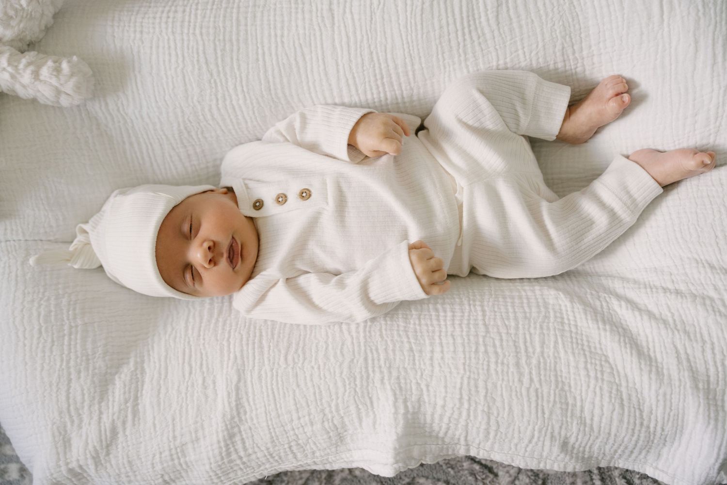 Newborn baby in cream outfit and hat sleeping peacefully on white bedding.