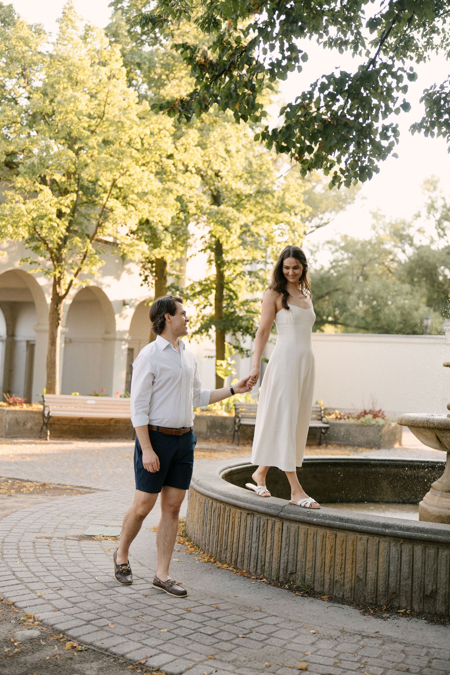 Woman in white dress standing on fountain edge holding mans hand in sunny park.
