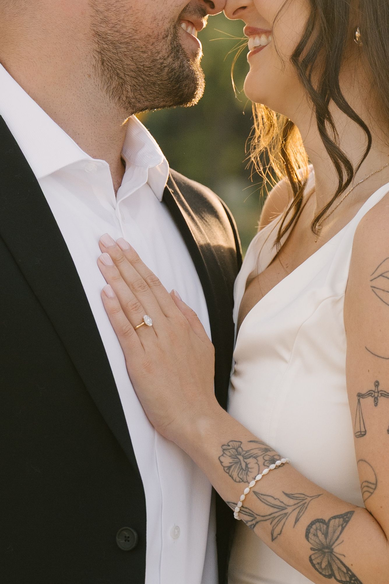 Couple nearly kissing at golden hour, woman showing diamond ring and tattoos on arm.