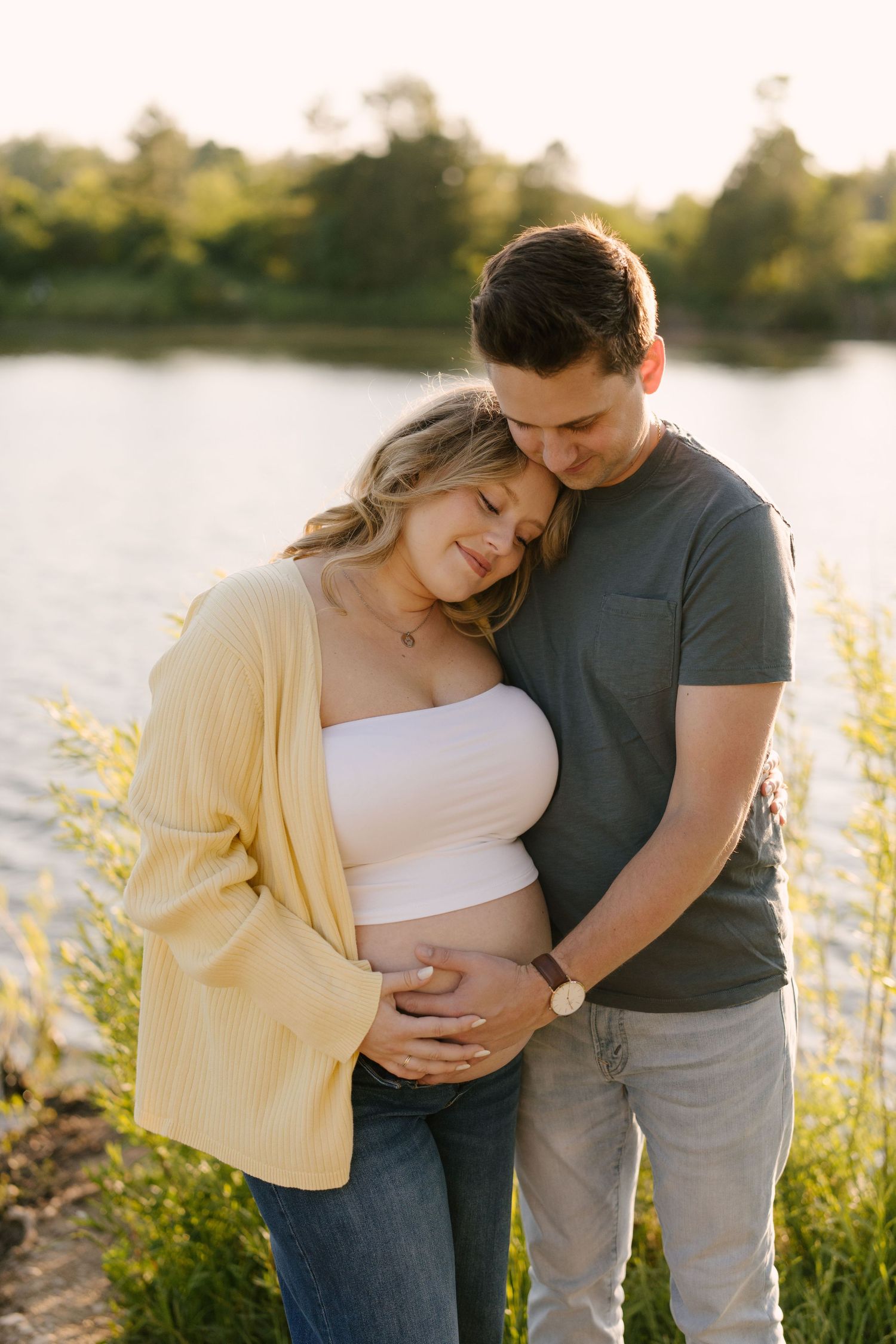 Couple tenderly embraces by a sunlit lake, both cradling the pregnant woman's baby bump at sunset.