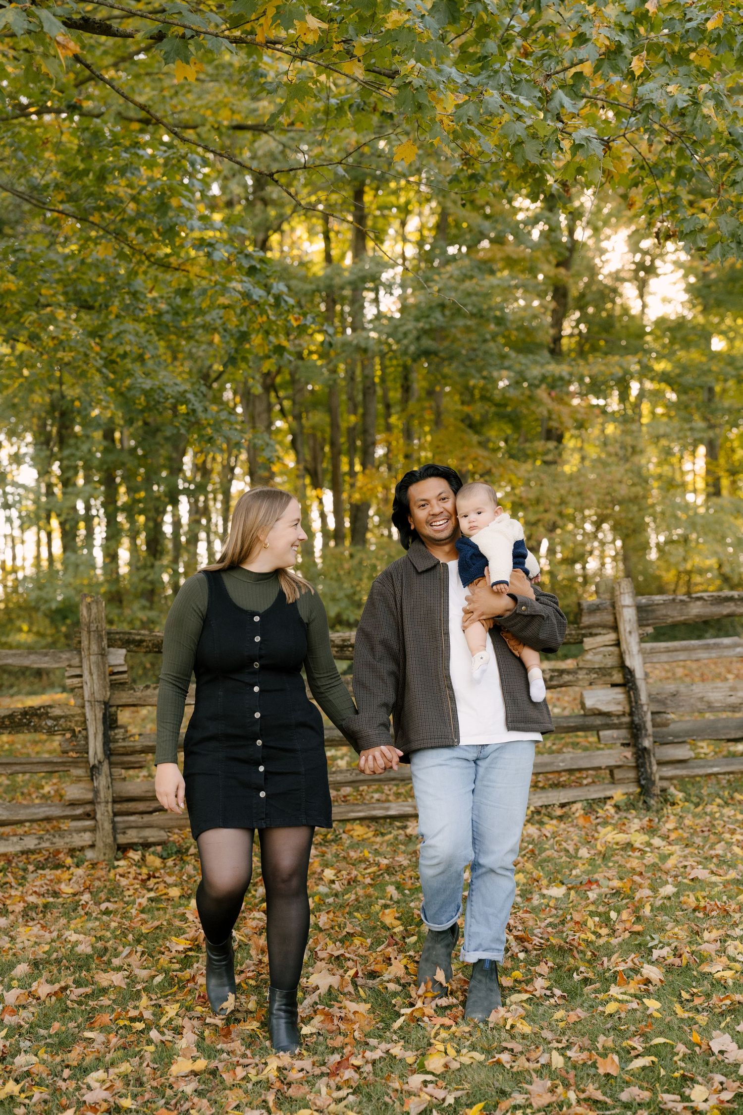 Happy interracial couple walking hand-in-hand in fall park, man carrying smiling baby.