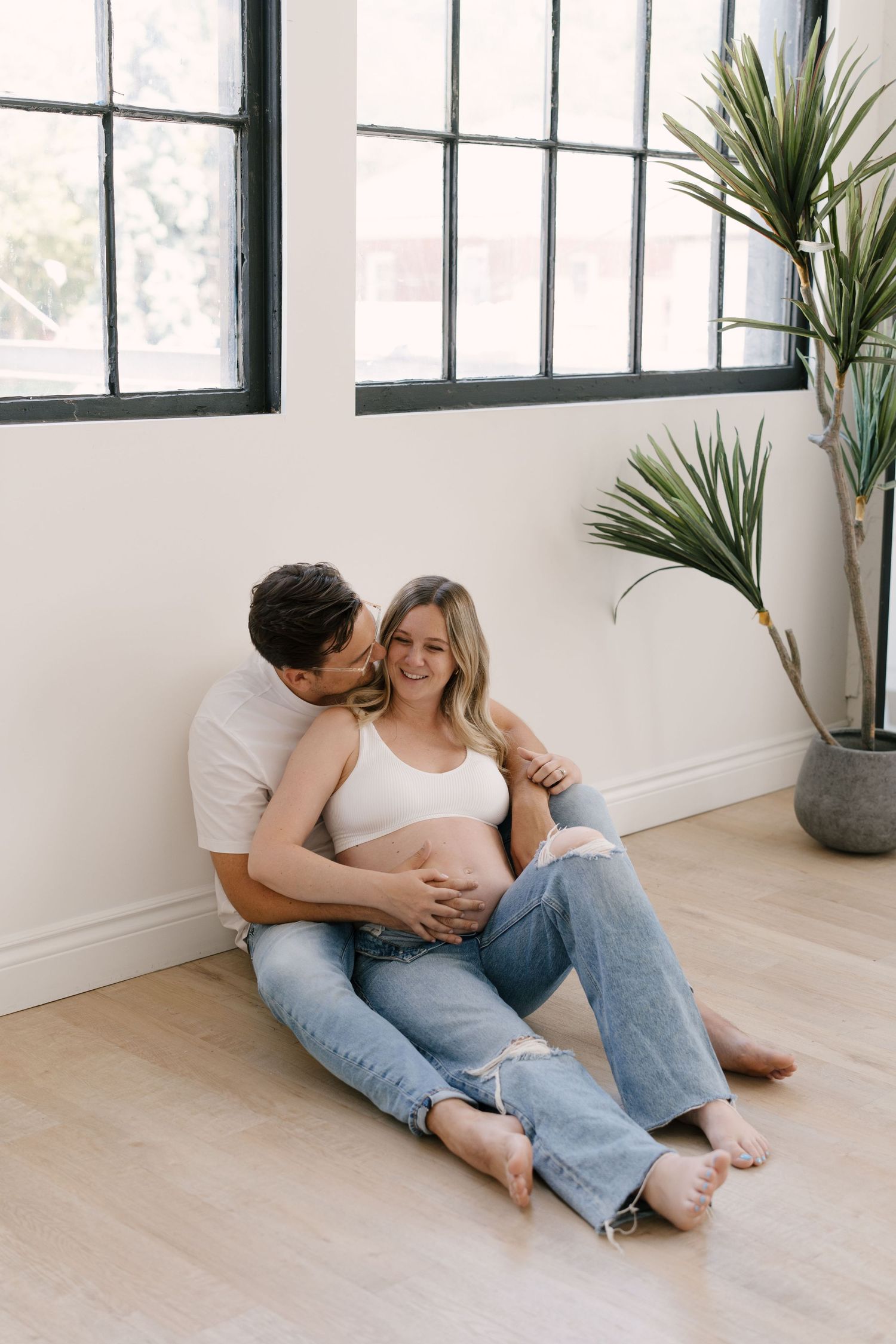 Couple sitting on floor, man embracing pregnant woman in white top and ripped jeans near large windows.