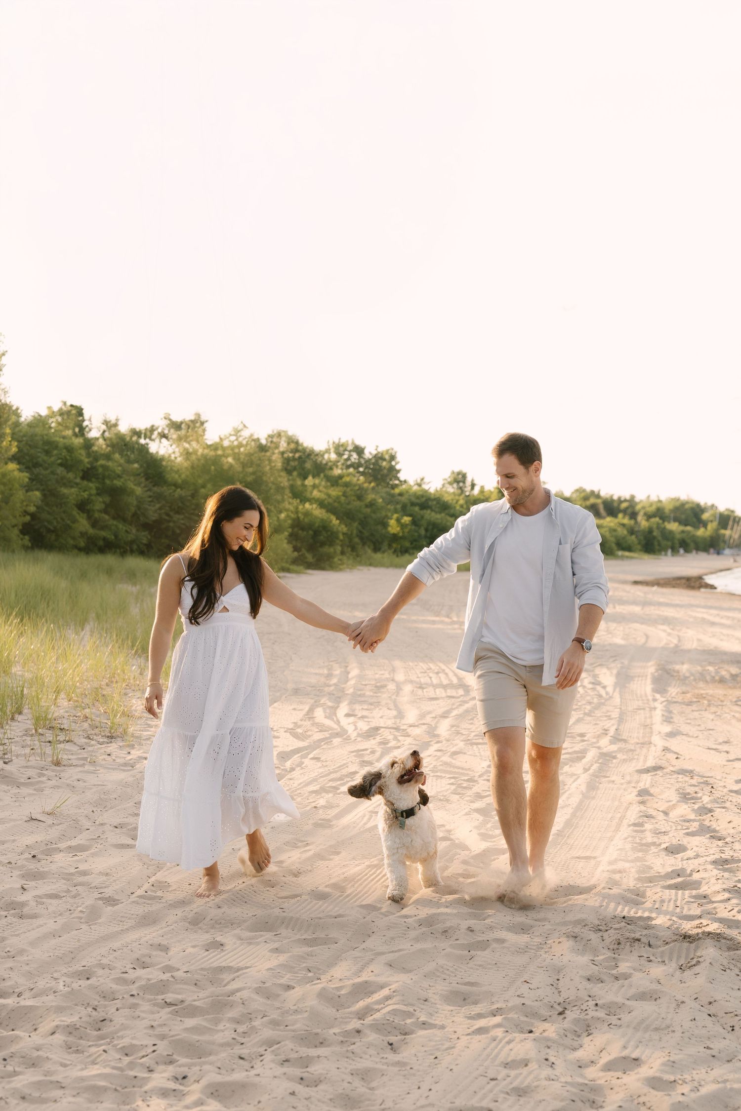 Couple walking hand in hand with small dog on sandy beach at sunset.