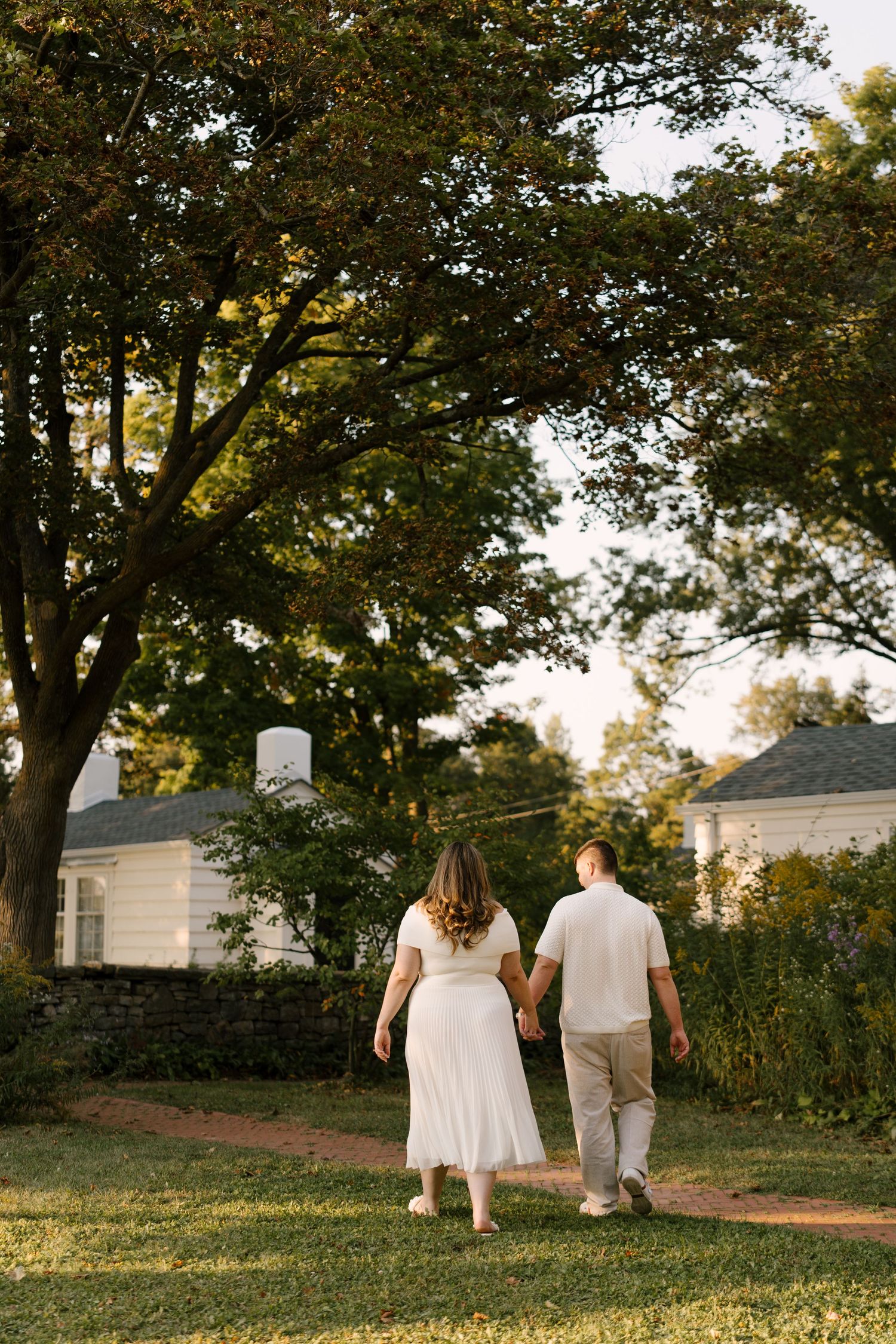 Couple walking hand-in-hand along a garden path toward white cottages beneath a canopy of trees.