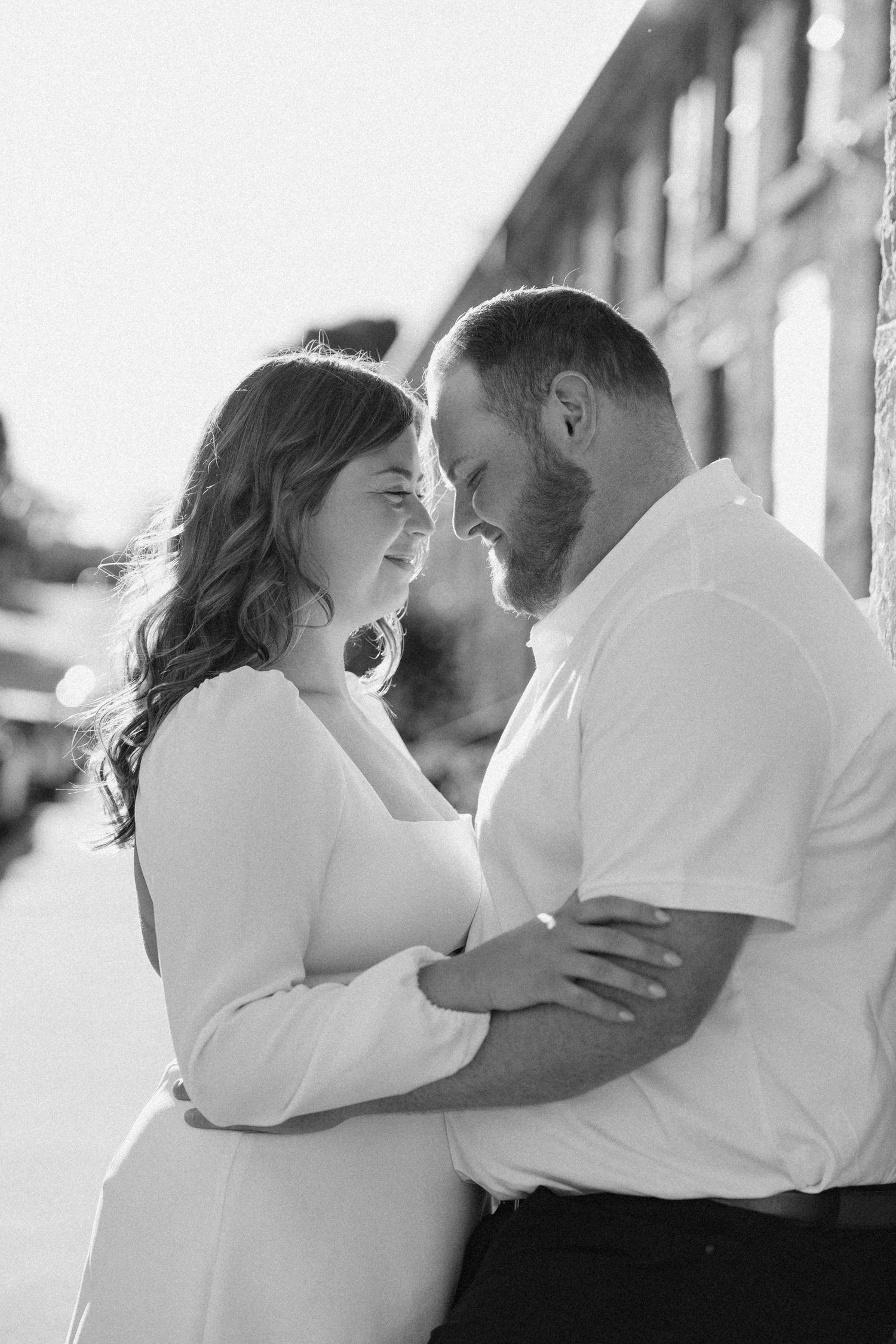 Black and white photo of couple embracing and smiling at each other near a building.